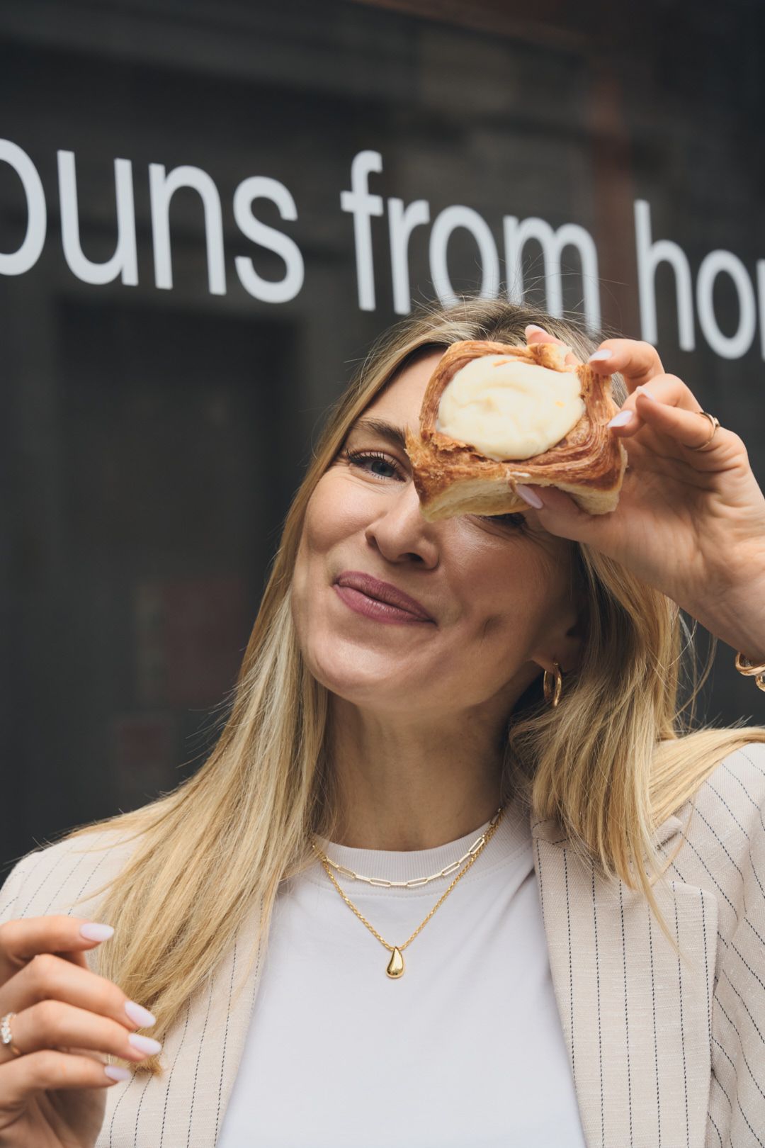 Person in white pinstripe outfit playfully holding a pastry or baked good in front of their face.