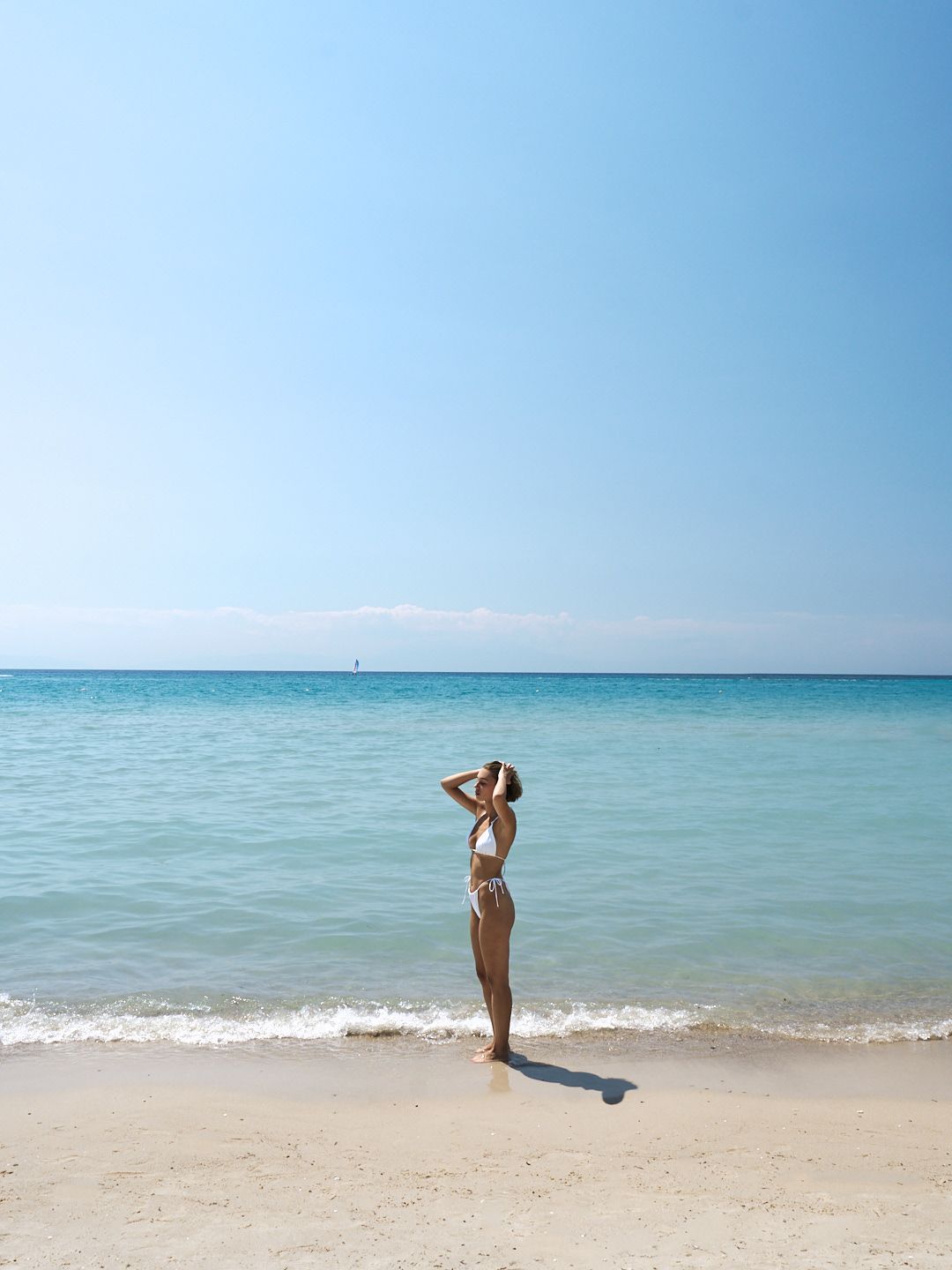 Woman in white bikini standing on sandy beach with calm turquoise ocean and blue sky.