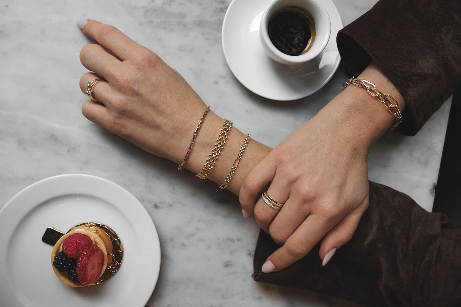 Delicate gold bracelets adorn a wrist next to a coffee cup and strawberry dessert on marble table.