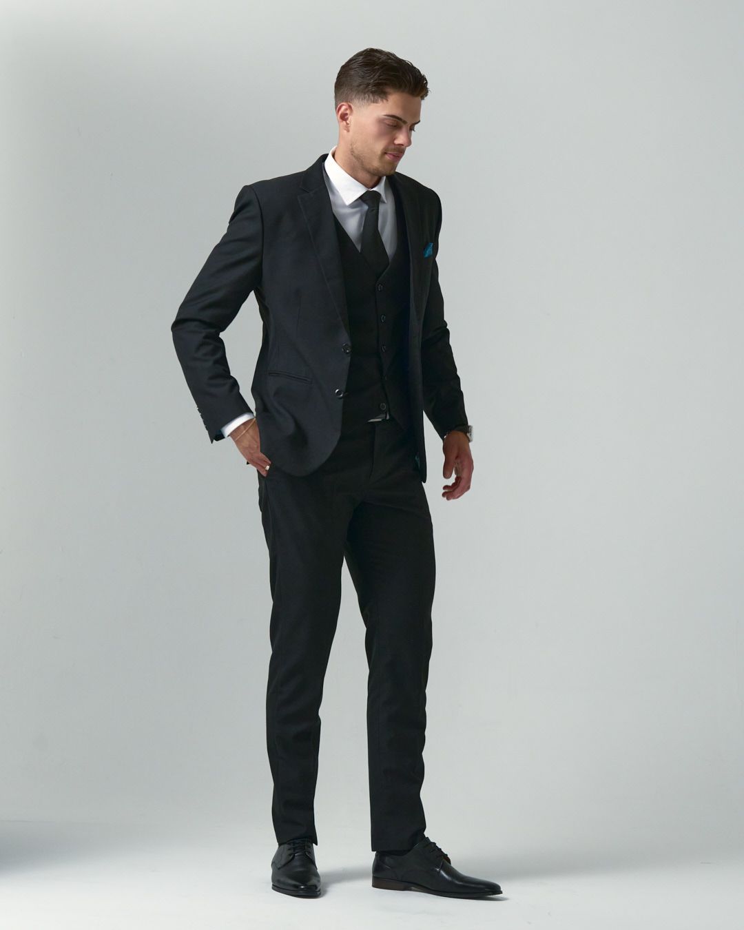 Well-dressed individual in black three-piece suit and tie poses against light gray background in professional stance.