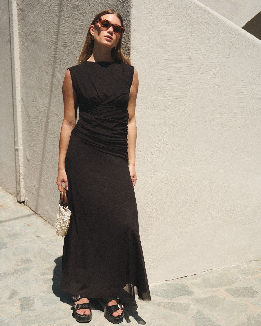 Woman in flowy black dress posing against whitewashed wall in a Mediterranean setting with sunglasses.