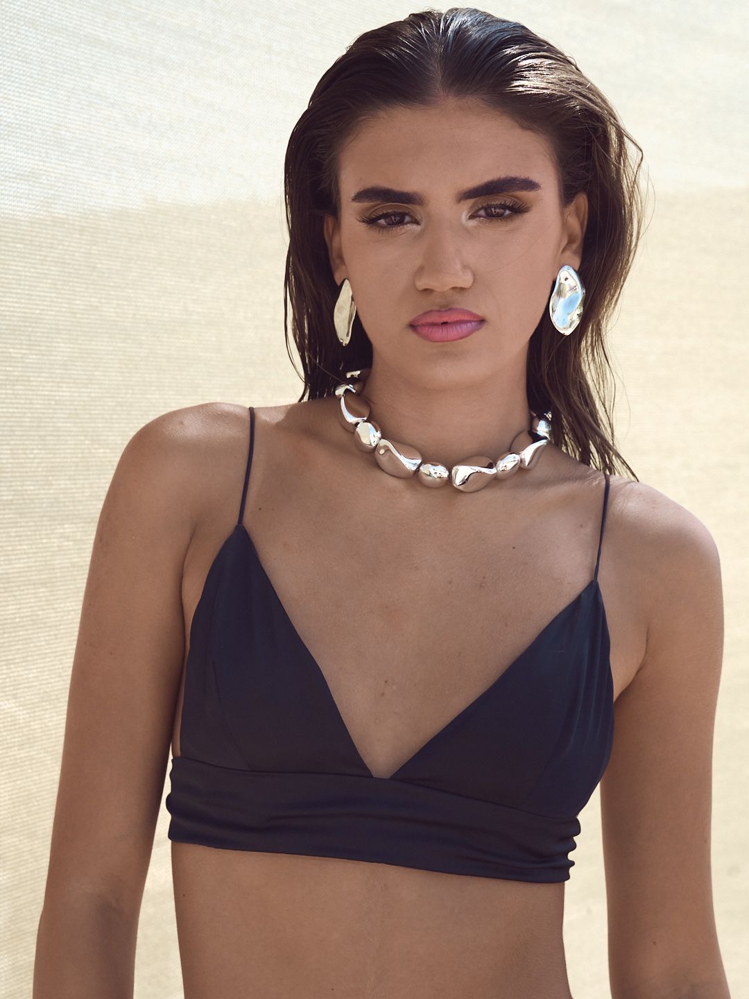 Woman in black bikini top with shell necklace and earrings posing on a sandy beach.