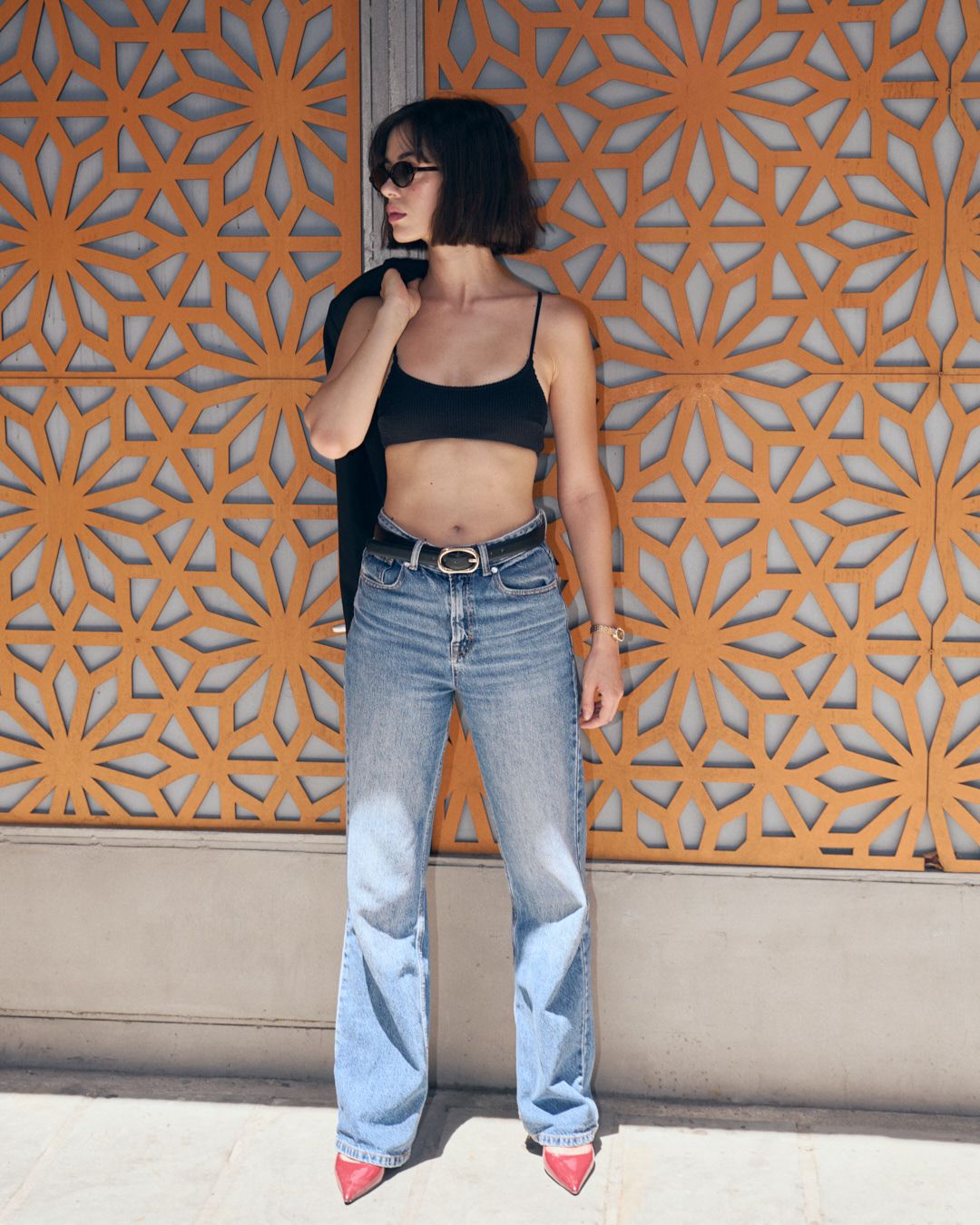 Person in retro outfit poses against orange floral wall backdrop wearing light-wash jeans, black top, and red heels.