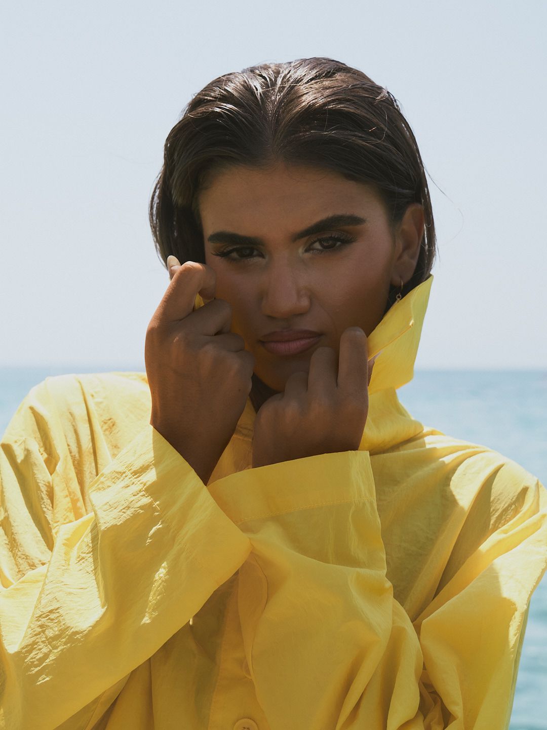 Young woman in yellow raincoat with wet hair posing against ocean backdrop on overcast day.