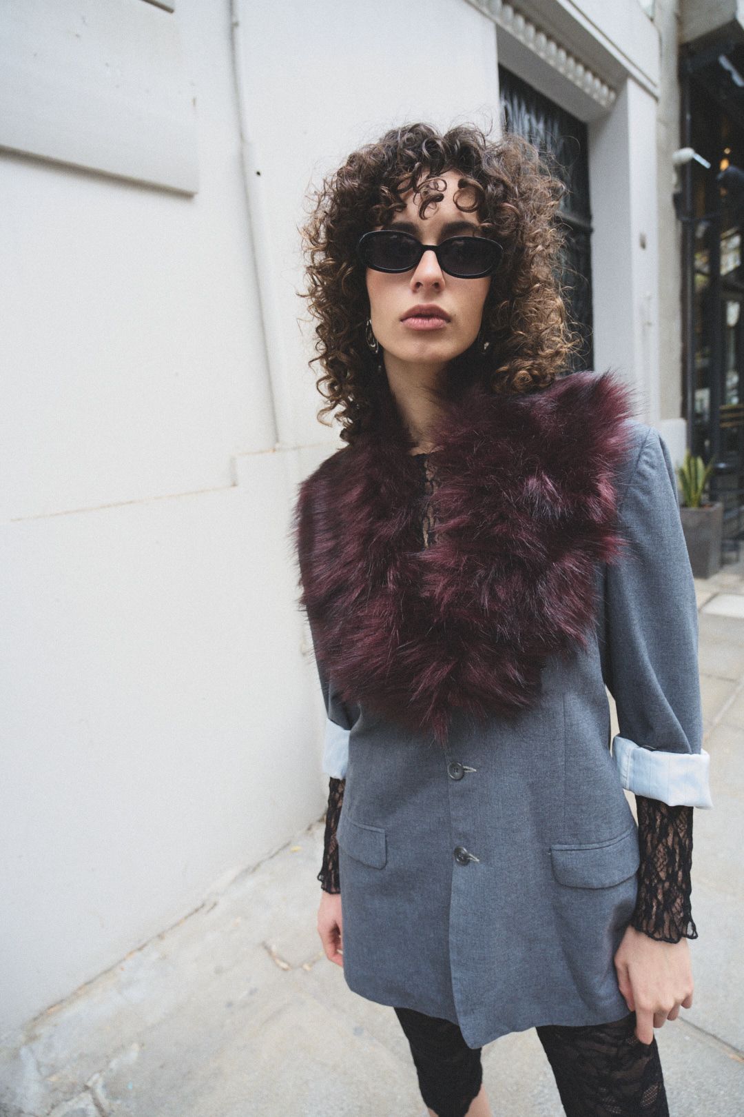 Fashion model wearing a gray blazer with burgundy fur collar and dark curly hair poses against white wall.