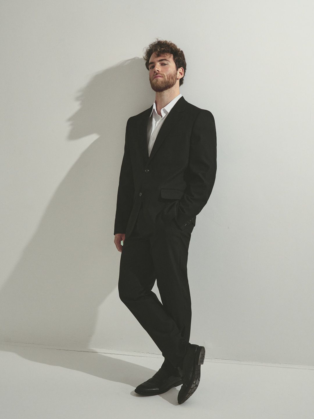 A person in a black suit and white shirt poses standing against a white wall casting a dramatic shadow.