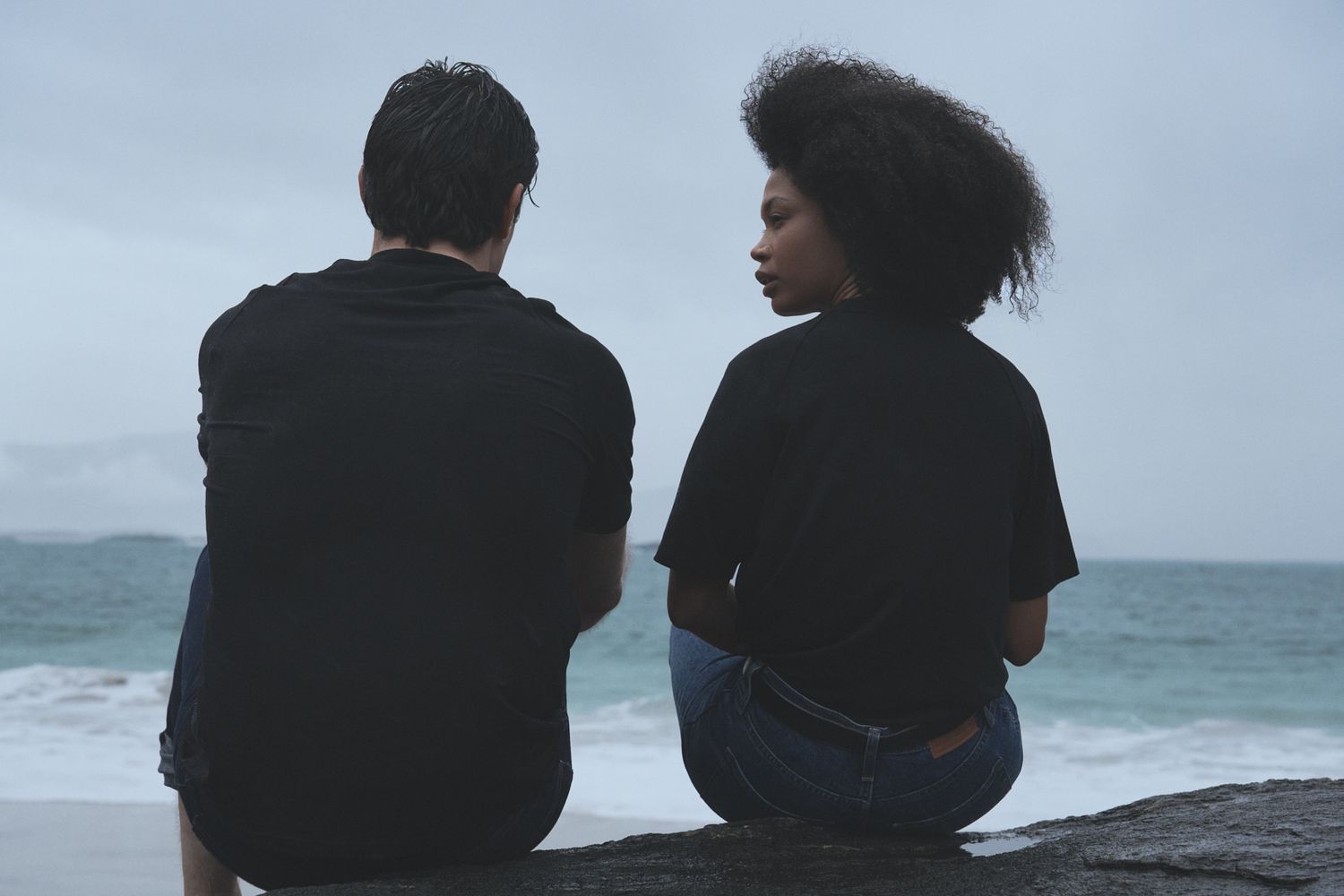 Two people sit together on a rocky cliff overlooking the ocean on a cloudy day.
