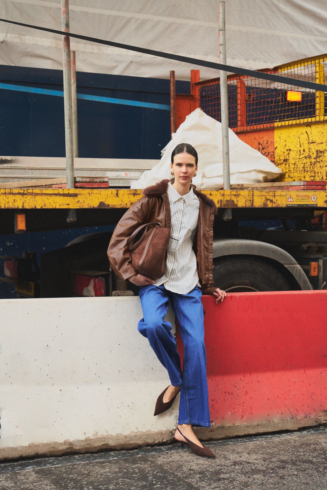A person in a brown jacket and blue pants sits casually on a red and white concrete barrier in an urban setting.