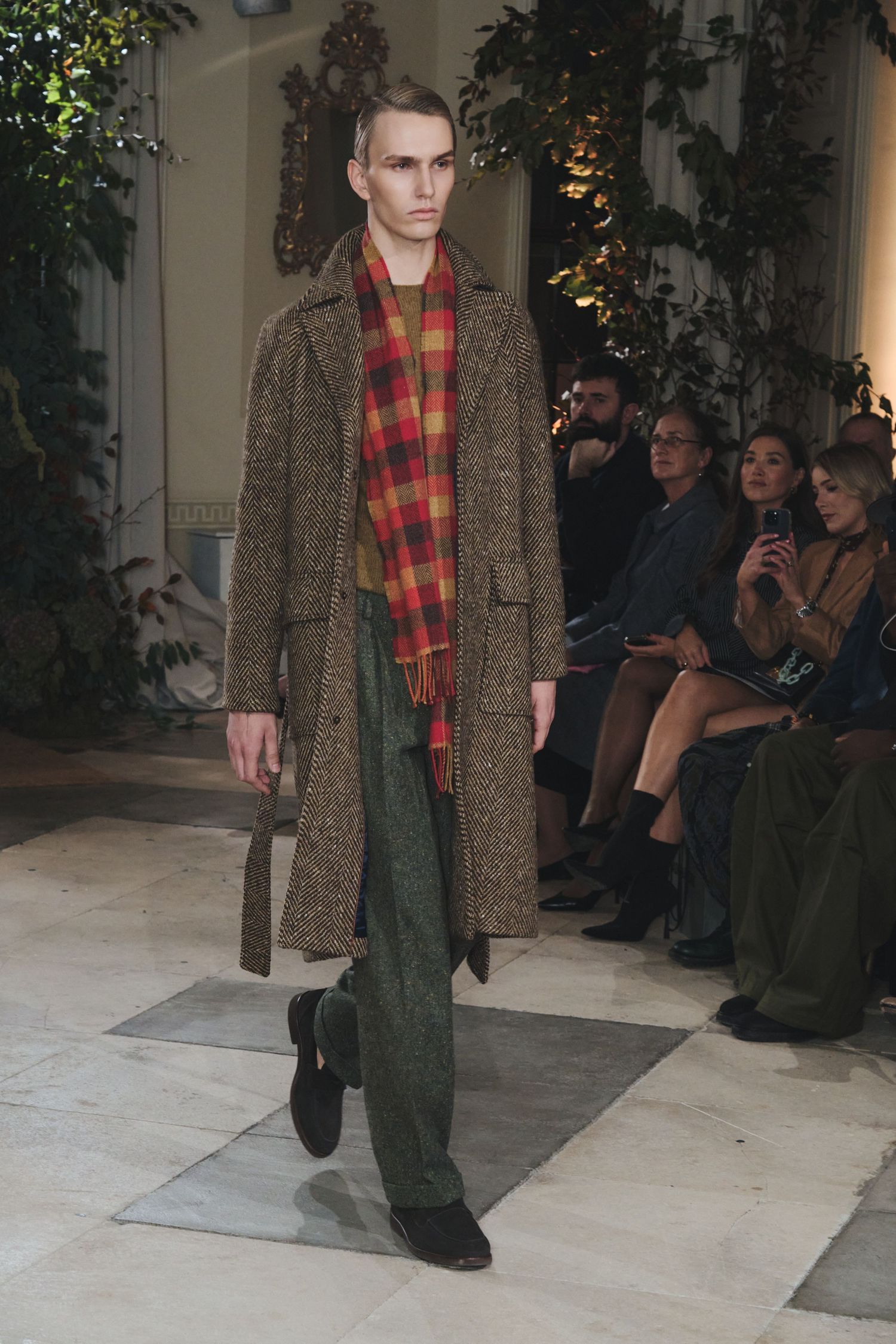 Model walks runway wearing brown tweed coat, plaid scarf and olive pants during fashion show presentation.
