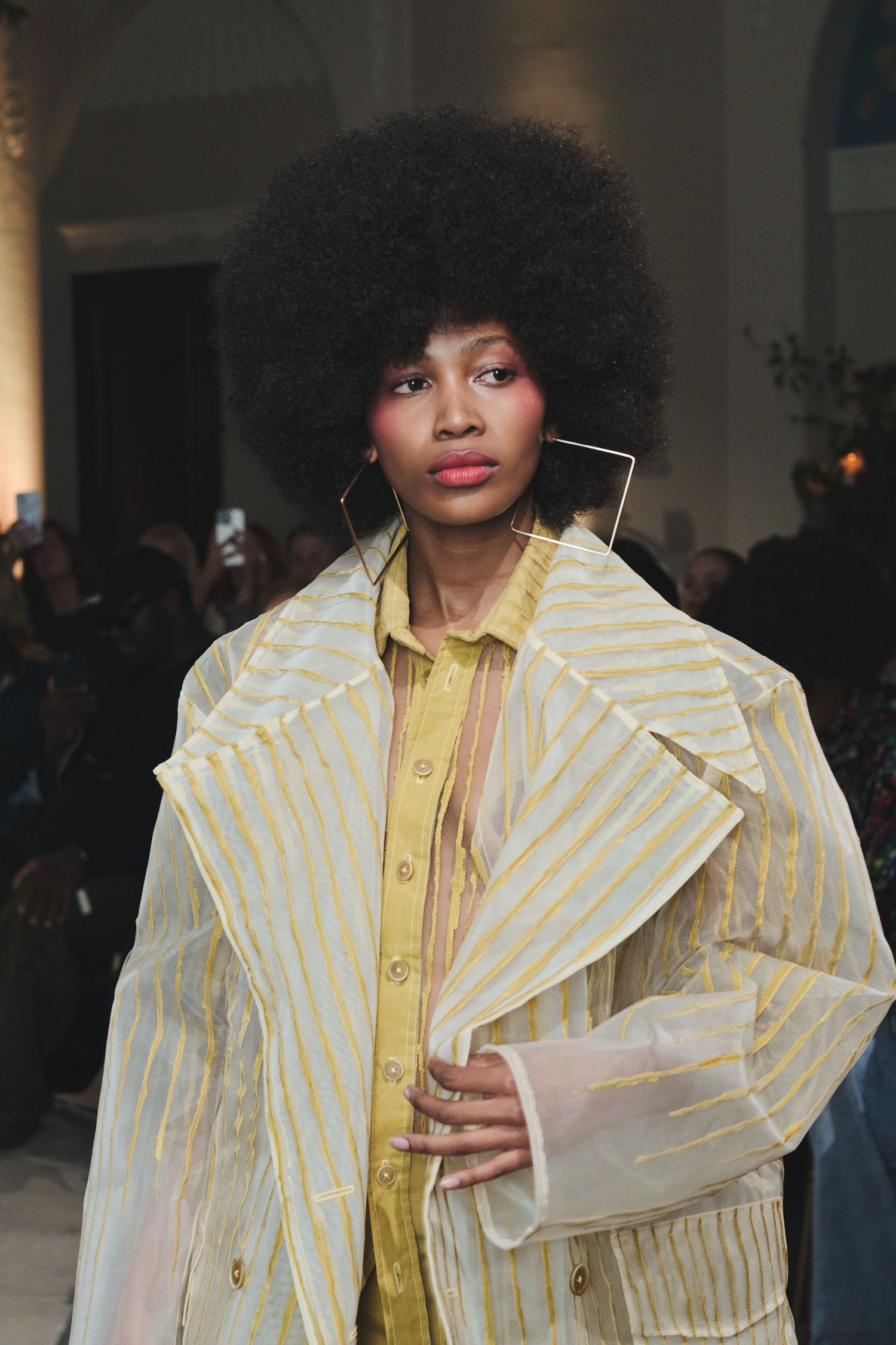 Fashion model walks runway in yellow striped designer coat with dramatic collar and matching yellow blouse.