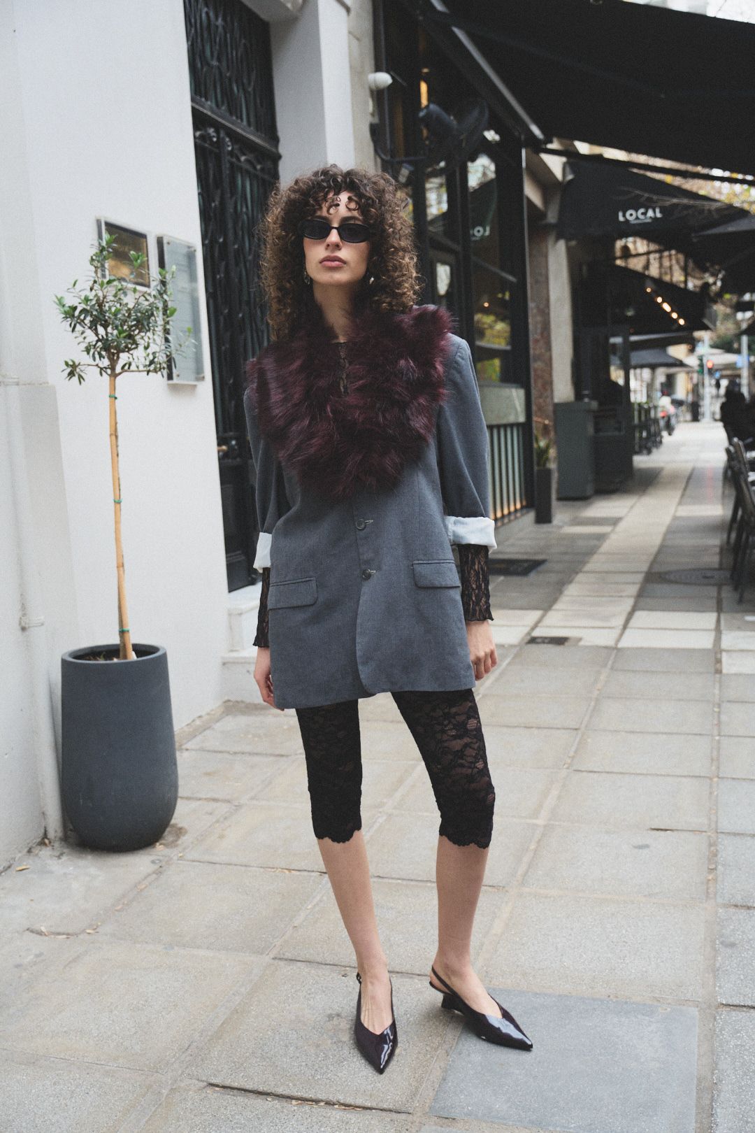 Fashionable person wearing grey blazer, burgundy fur collar, lace capri pants and sunglasses poses on city sidewalk.