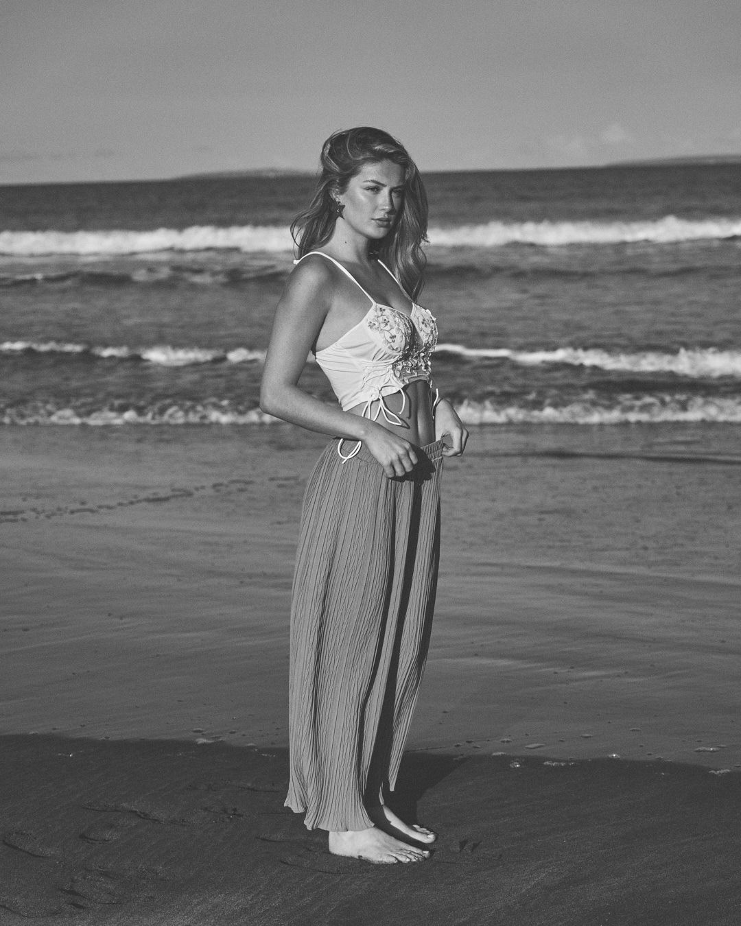 Black and white beach portrait of woman in flowing maxi dress with waves in background.