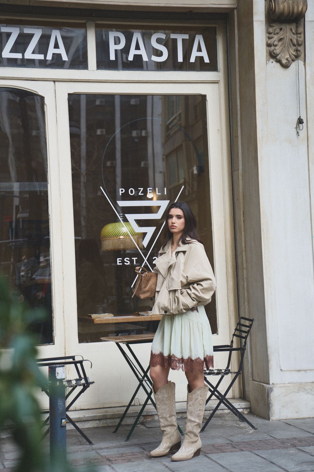 Woman in beige coat and boots poses outside a Pizza and Pasta restaurant storefront with black folding chairs.