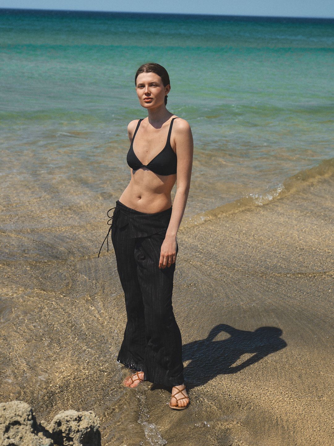 Woman in black bikini top and wrap skirt standing on sandy beach with turquoise ocean water behind her.