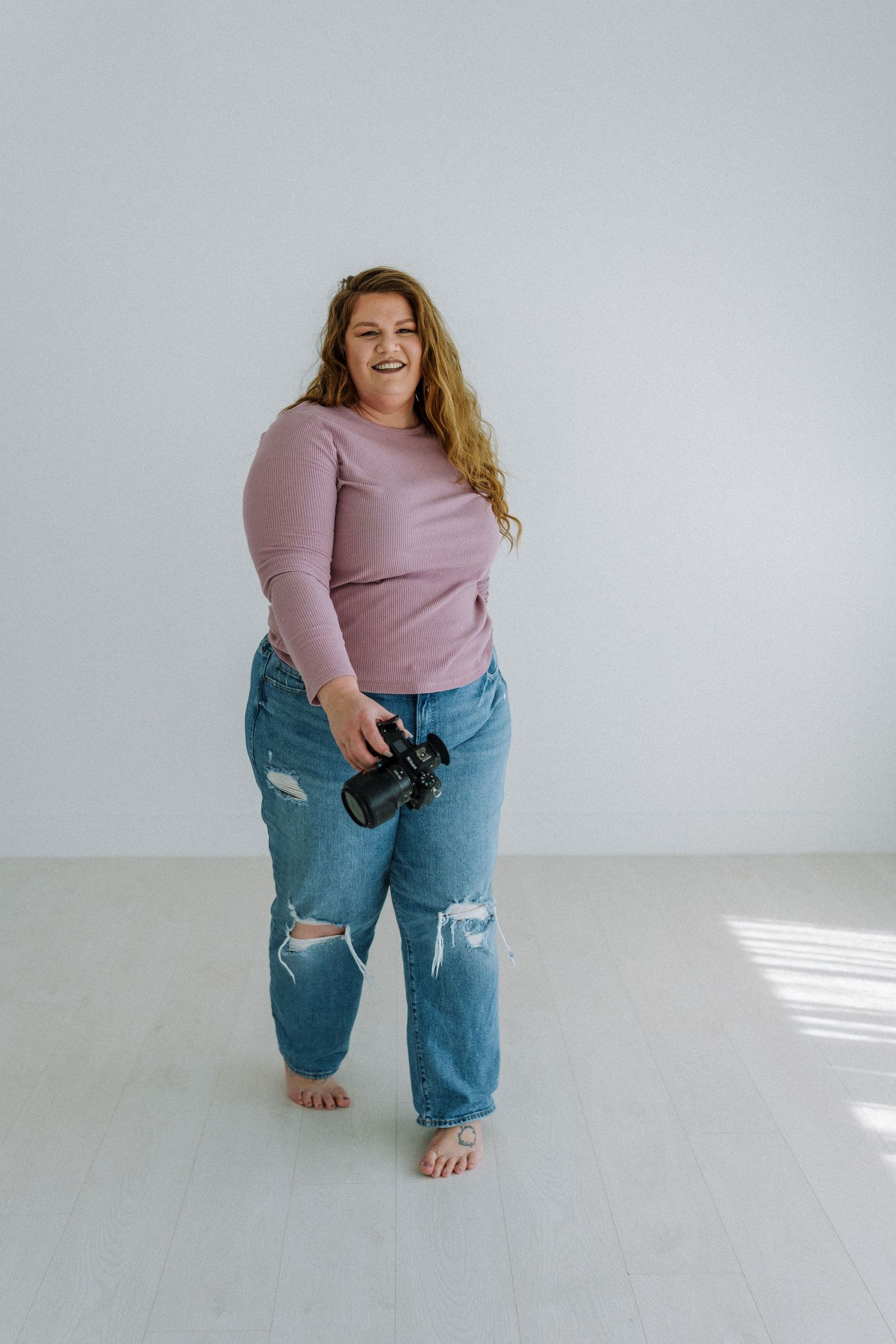 Woman in pink sweater and ripped jeans holding camera in bright studio setting.