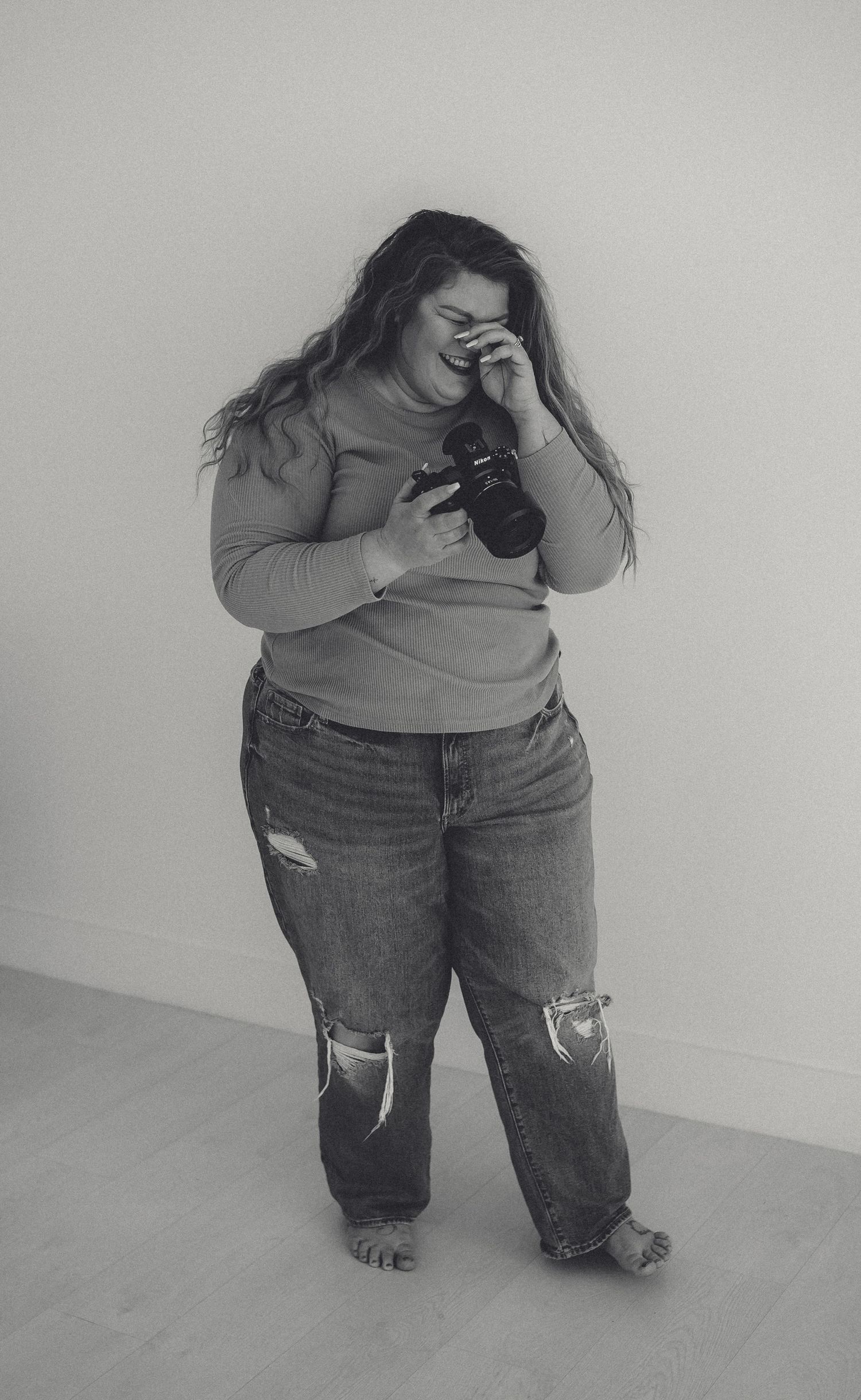 Woman in casual attire laughing while holding camera in minimalist studio setting.