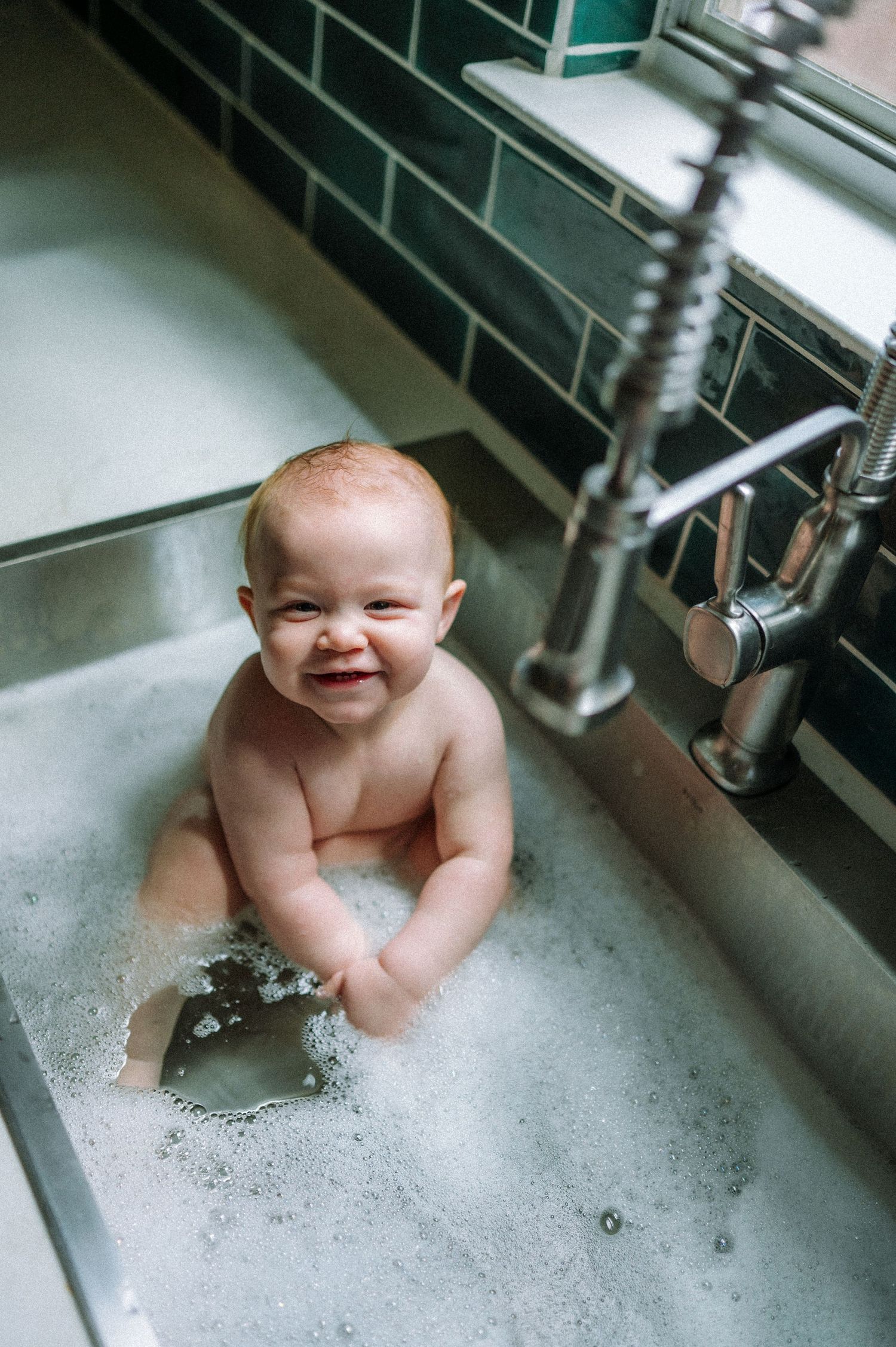 Happy baby playing in kitchen sink bath with bubbles.