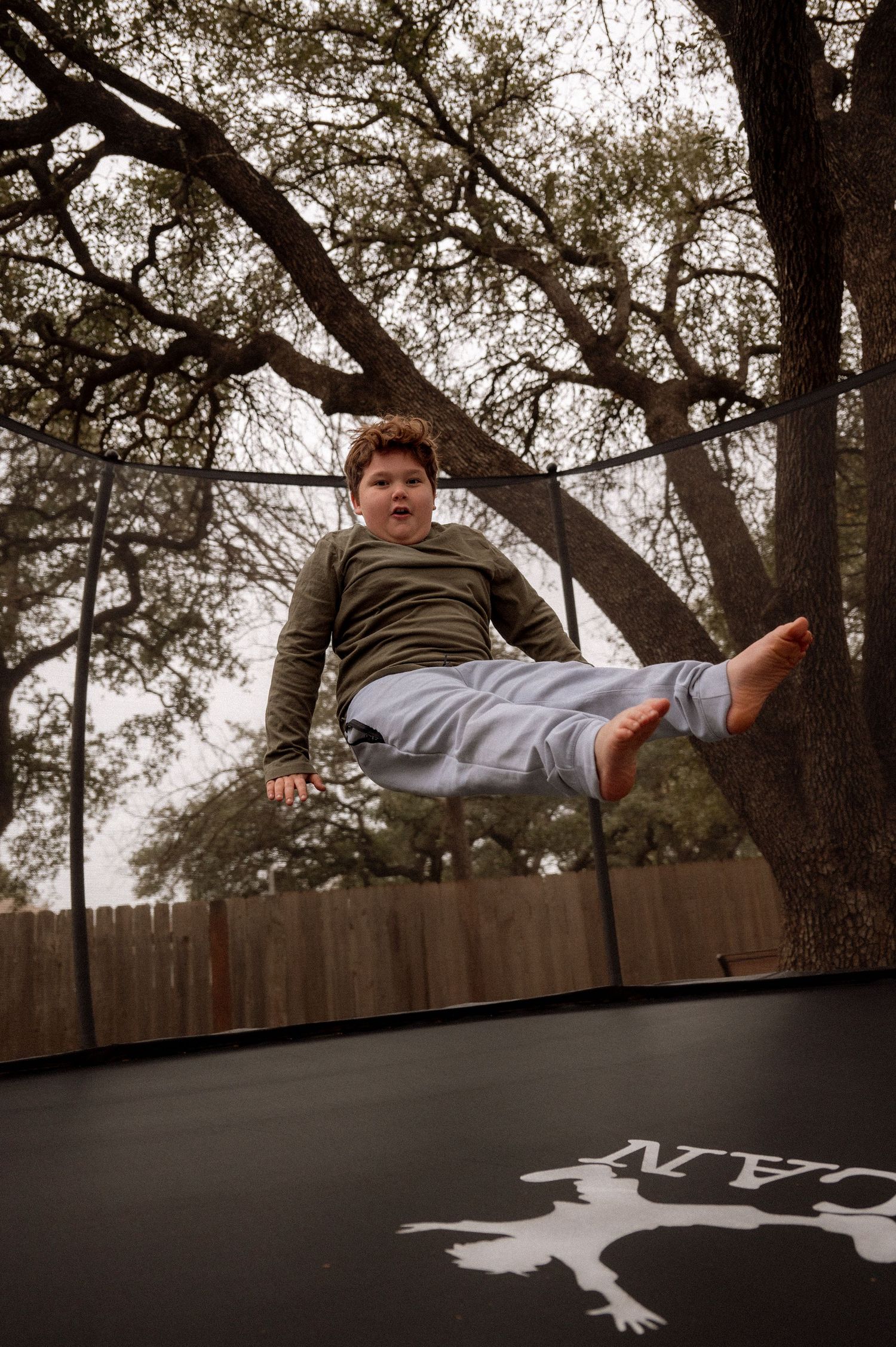 Child mid-jump on a backyard trampoline with tall trees in background.