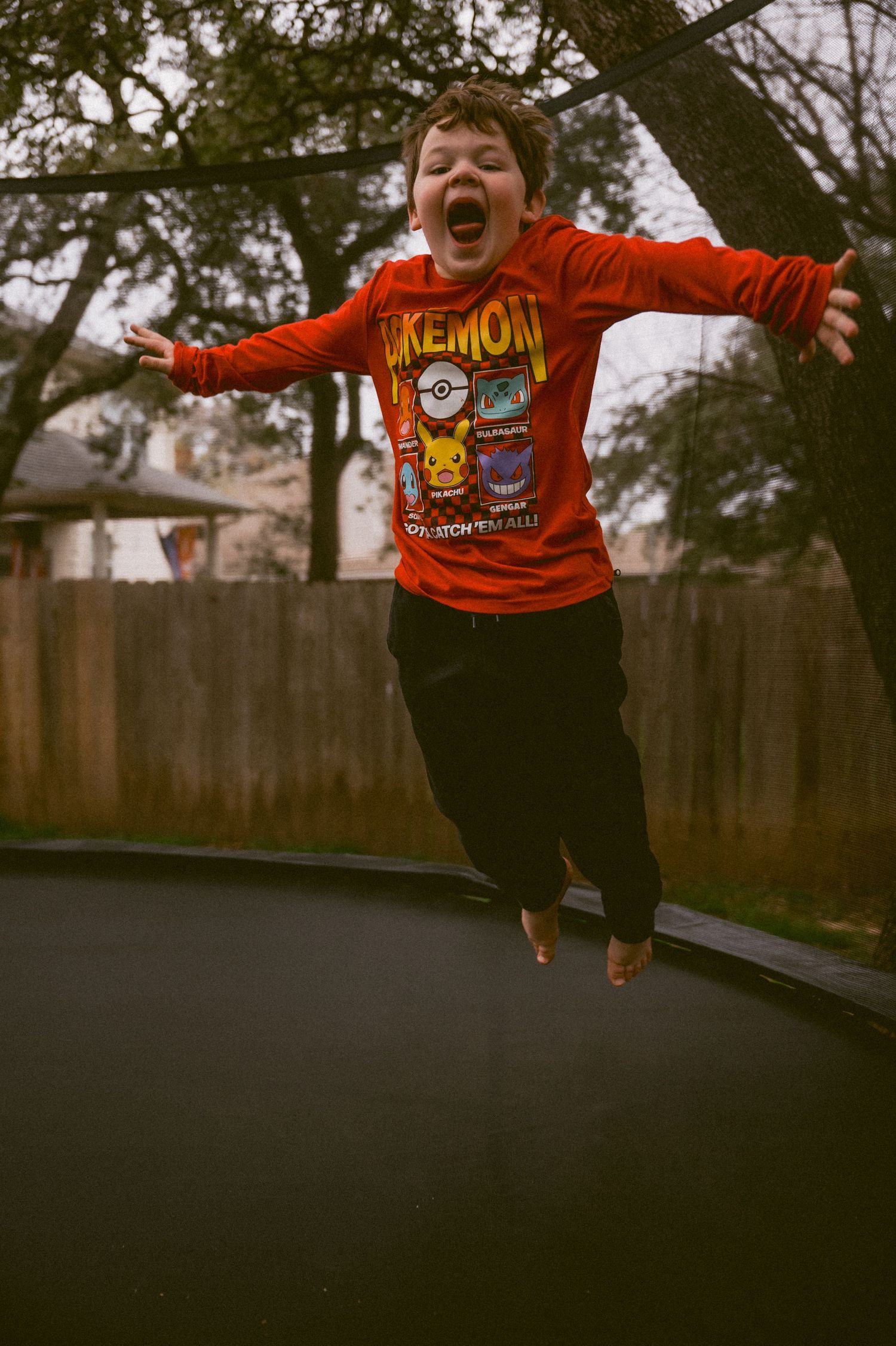 Child jumping with excitement on a backyard trampoline wearing a red Pokemon shirt.