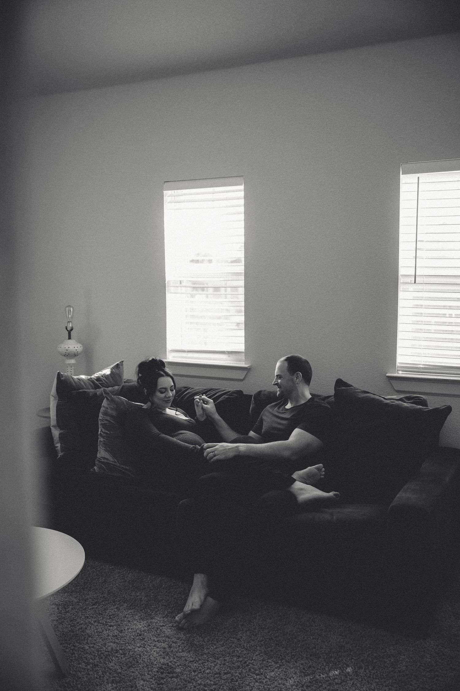 A black and white photo of two figures relaxing together on a couch in a living room with natural window light.