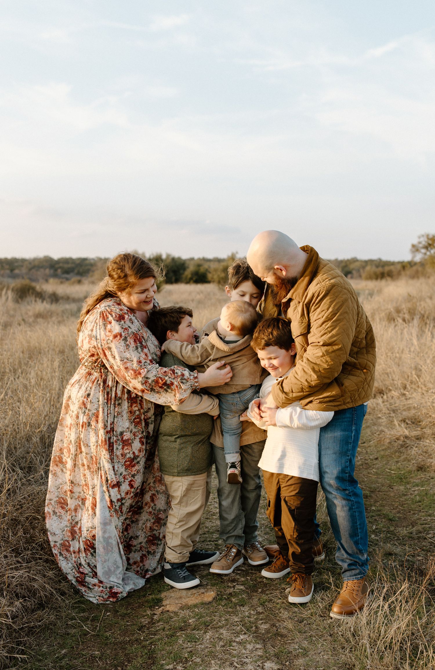 A family shares an intimate group hug outdoors in a grassy field during golden hour.