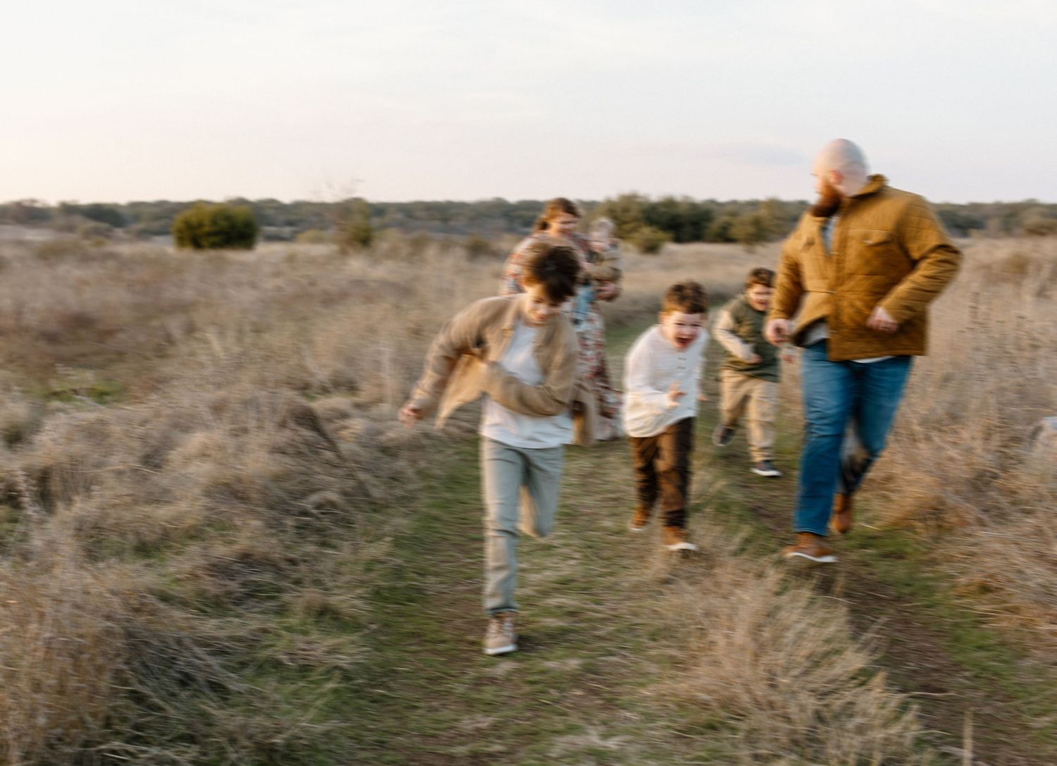 A family runs together along a nature trail through dried grass at sunset.