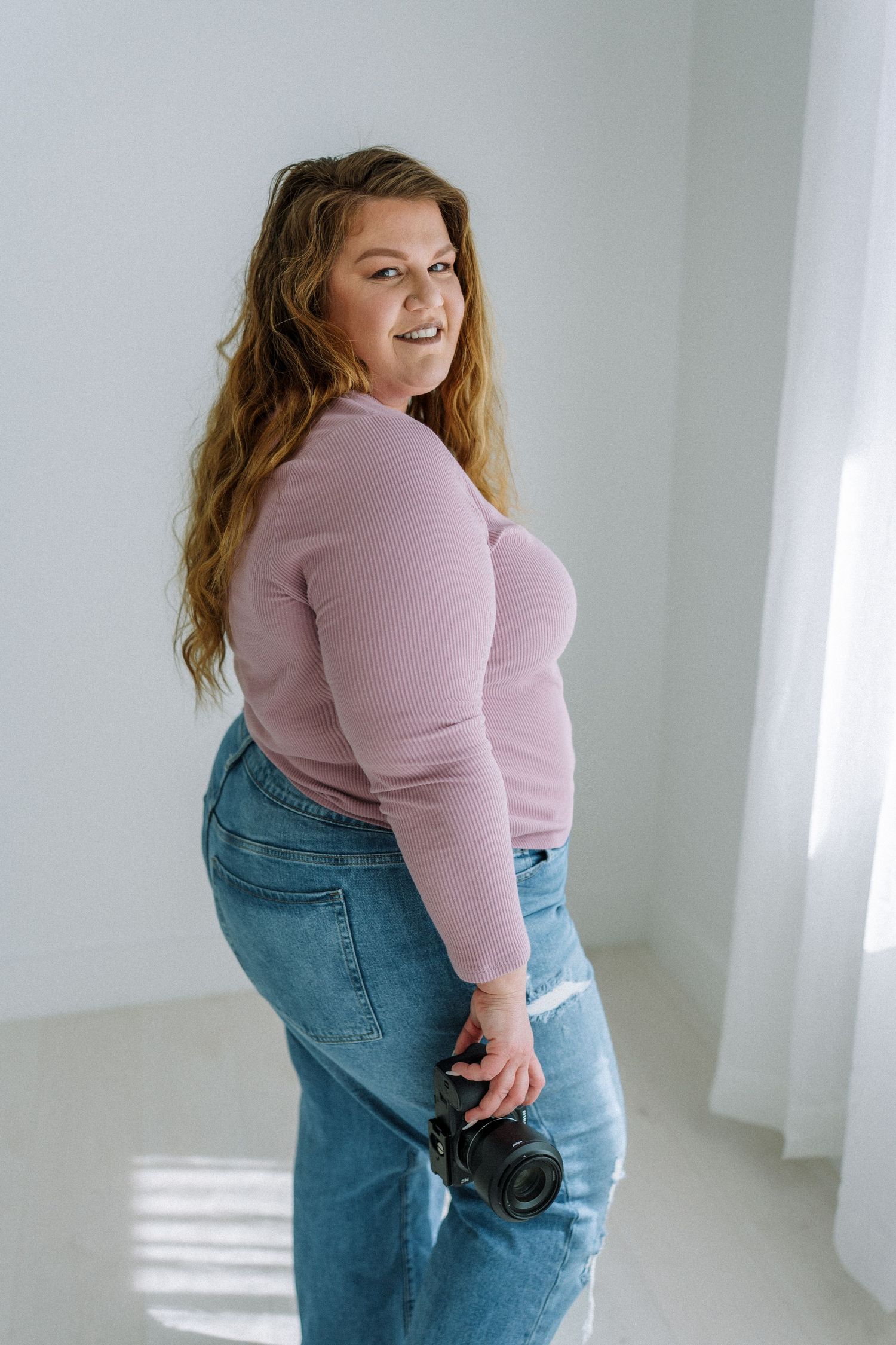 Woman in pink sweater and jeans poses with camera in bright studio setting.