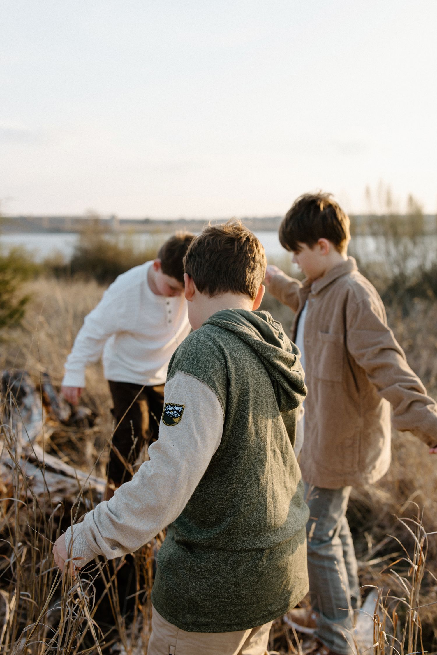Three people explore together in tall grass during golden hour near a lake.
