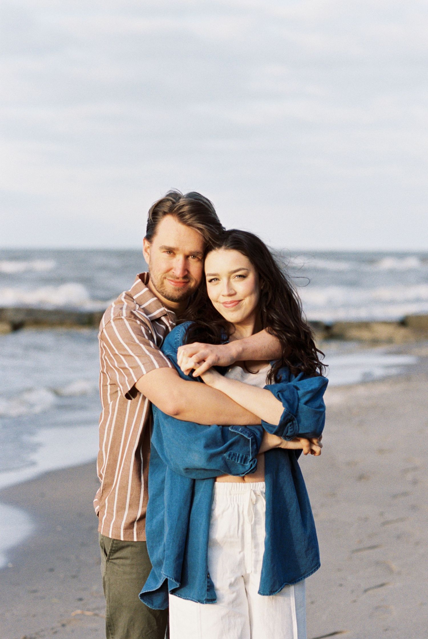 A couple embraces while standing on a beach with ocean waves in the background on a cloudy day.