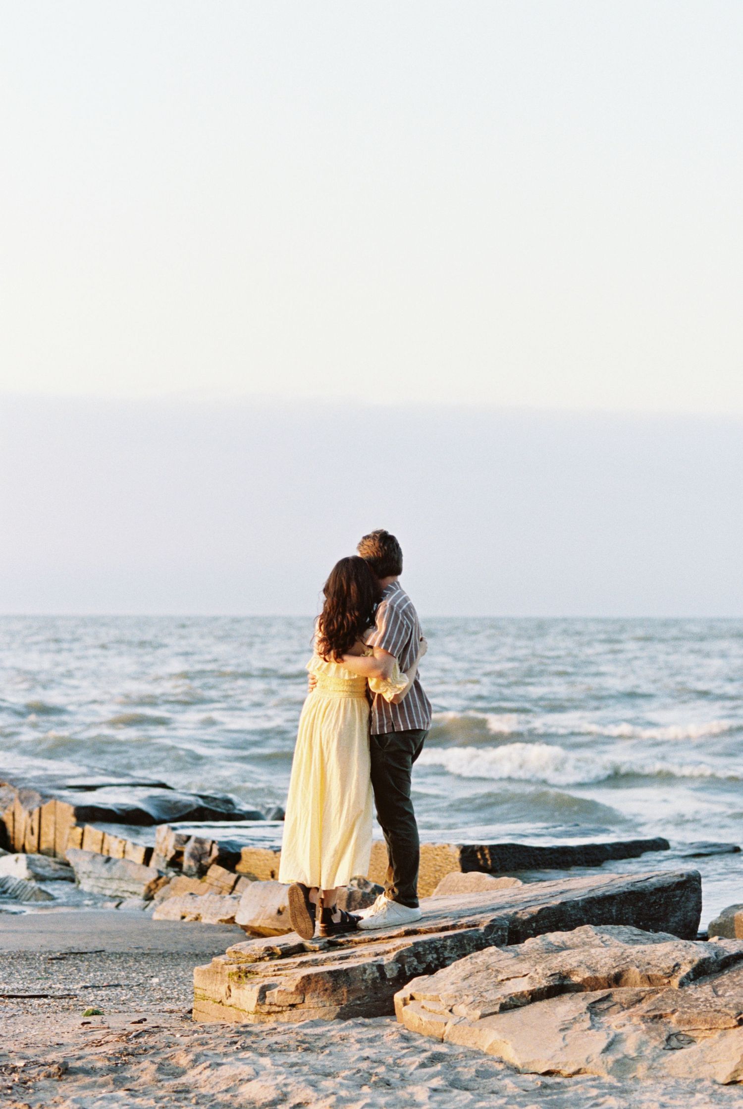 A couple embraces while standing on rocky coastal outcrop at sunset overlooking the ocean waves.