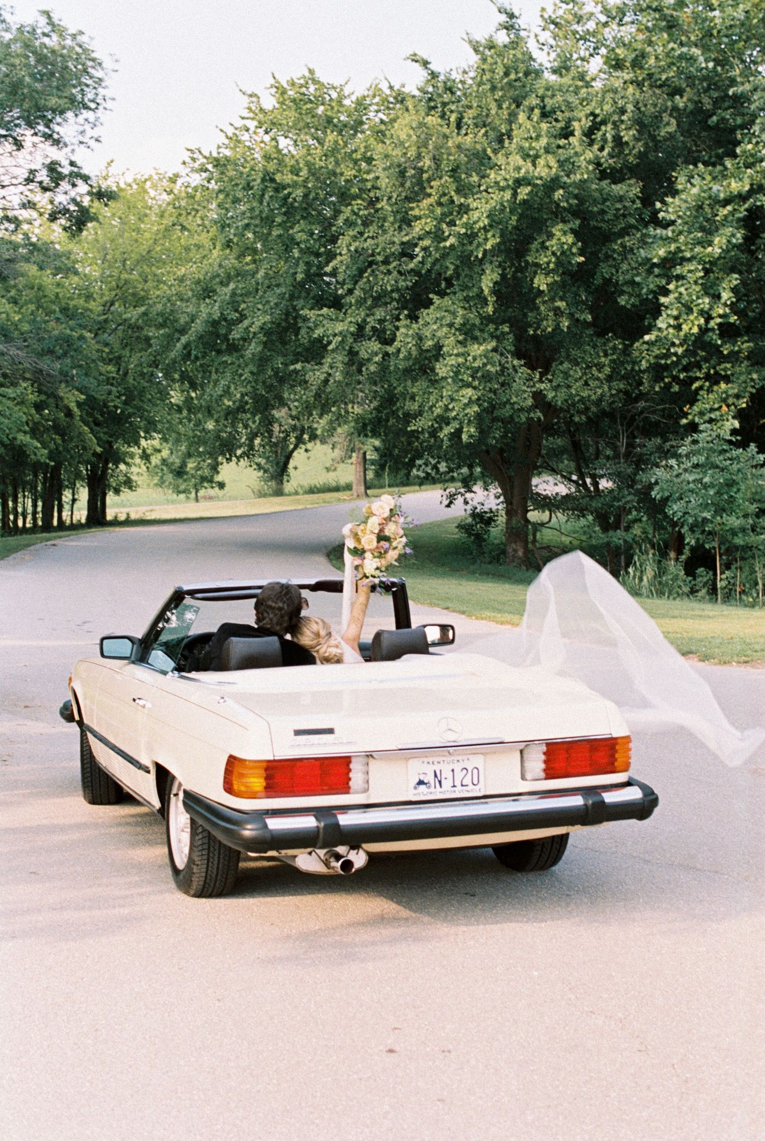 Classic white convertible Mercedes decorated with wedding flowers driving down a tree-lined road. Kirstie Whitaker Photography