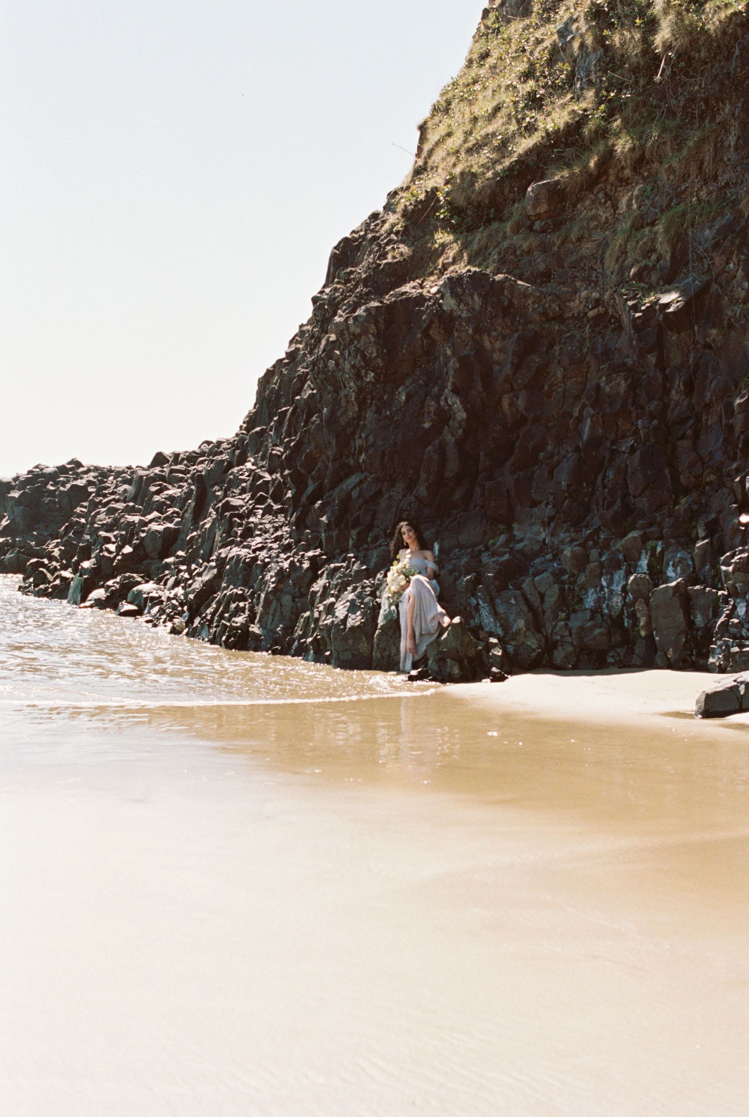 Figure in white dress stands on a sandy beach against dramatic rocky cliffs by the ocean. Kirstie Whitaker Photography