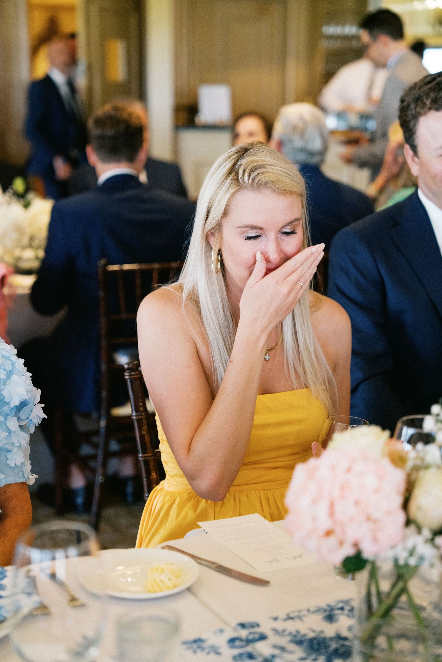 A wedding guest in a yellow dress laughs during a reception dinner, covering her mouth in genuine amusement.