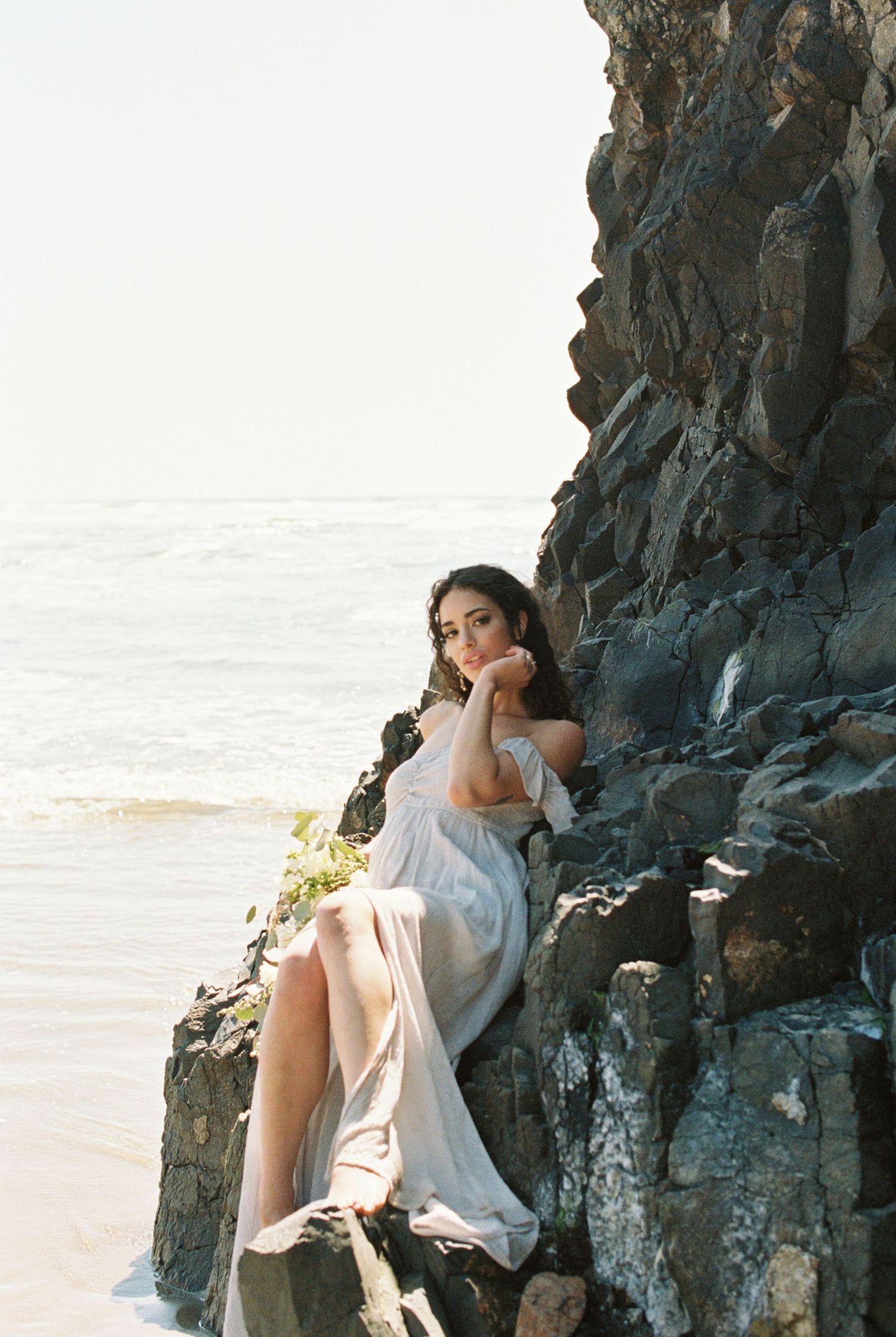 A figure in a flowing white dress poses elegantly against dramatic coastal cliffs at sunset by the ocean.