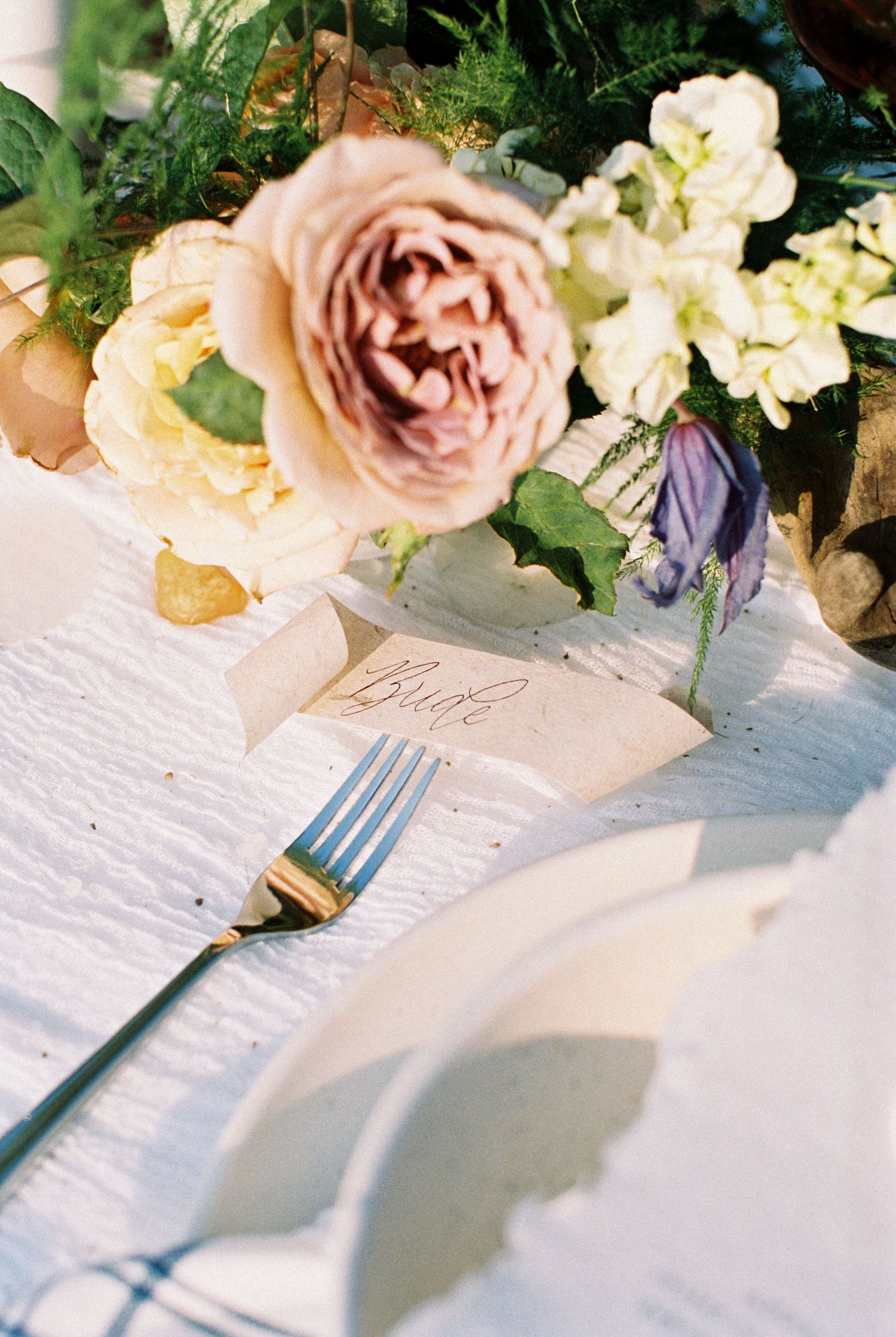 A delicate pink garden rose and white flowers in a vase beside a place setting with silverware on a white tablecloth.