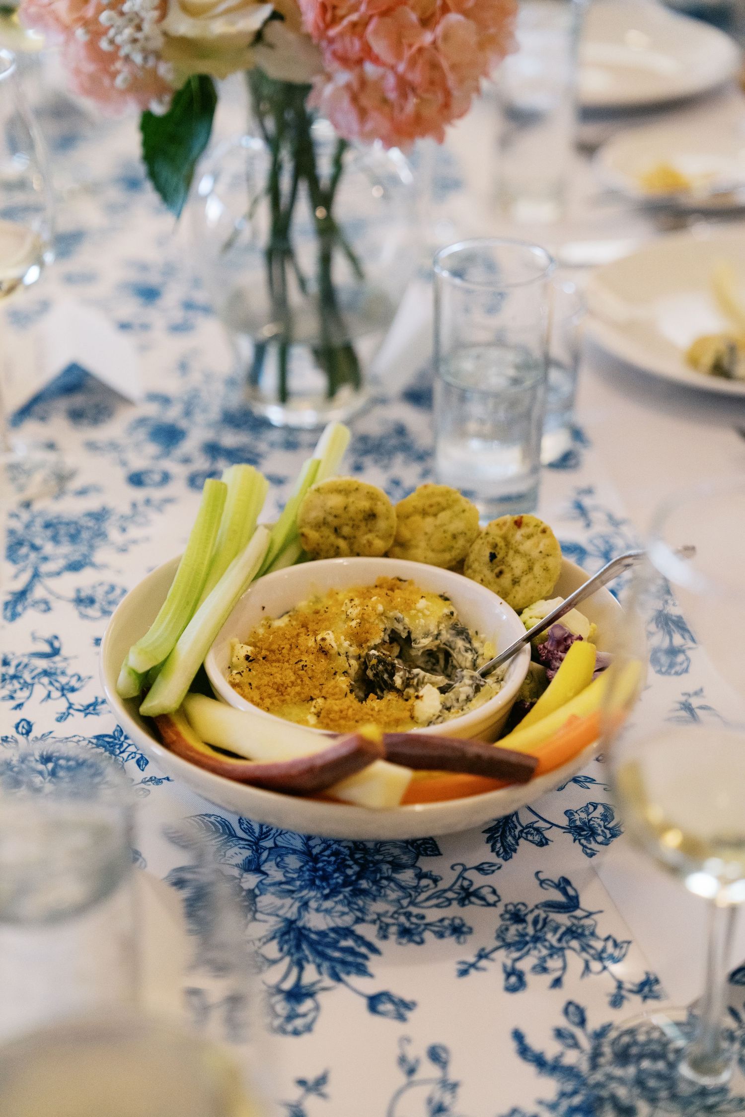Bowl of fresh vegetables and dip served on blue and white patterned tablecloth with pink flowers in vase.