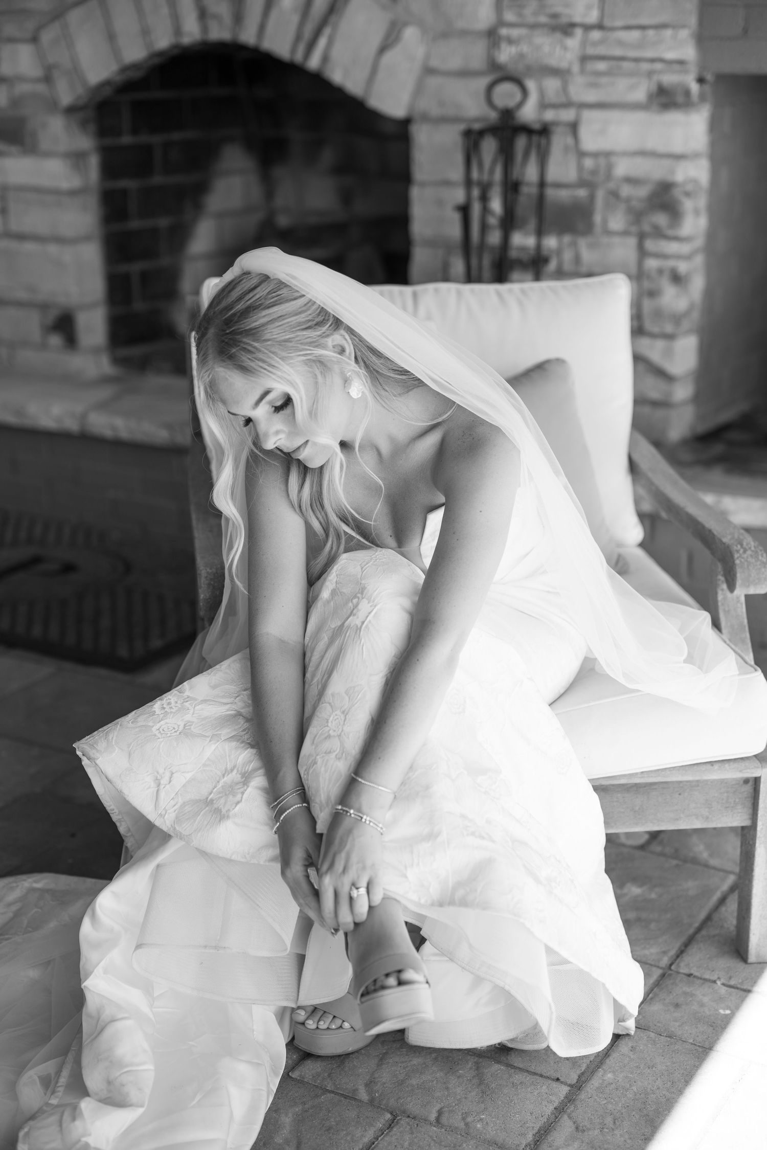 A bride in a flowing white wedding dress adjusts her shoe while sitting on stone steps in a black and white photograph.