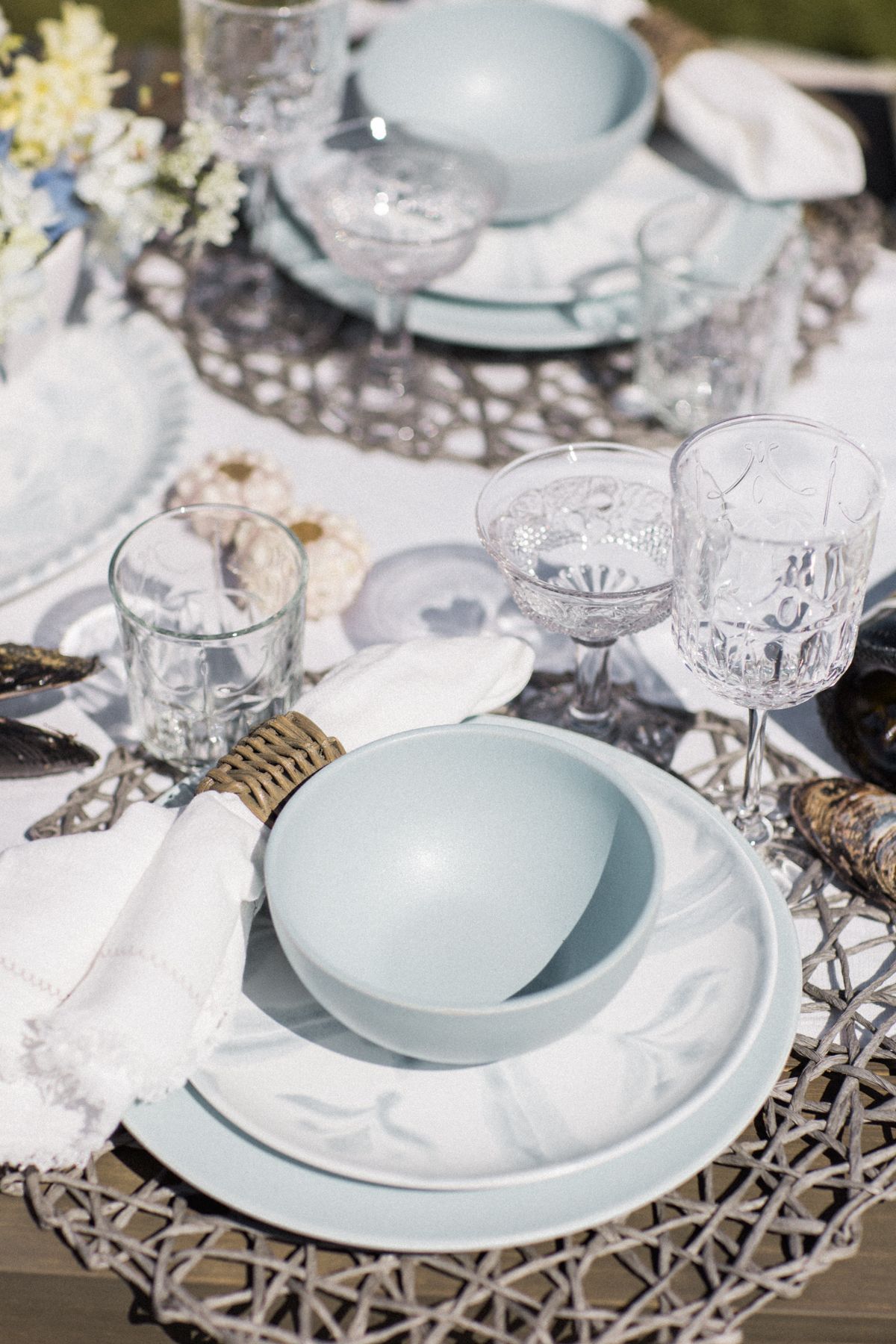 Elegant table setting with pale blue plates, crystal glassware, and woven placemats on a rustic wooden surface. Kirstie Whitaker Photography