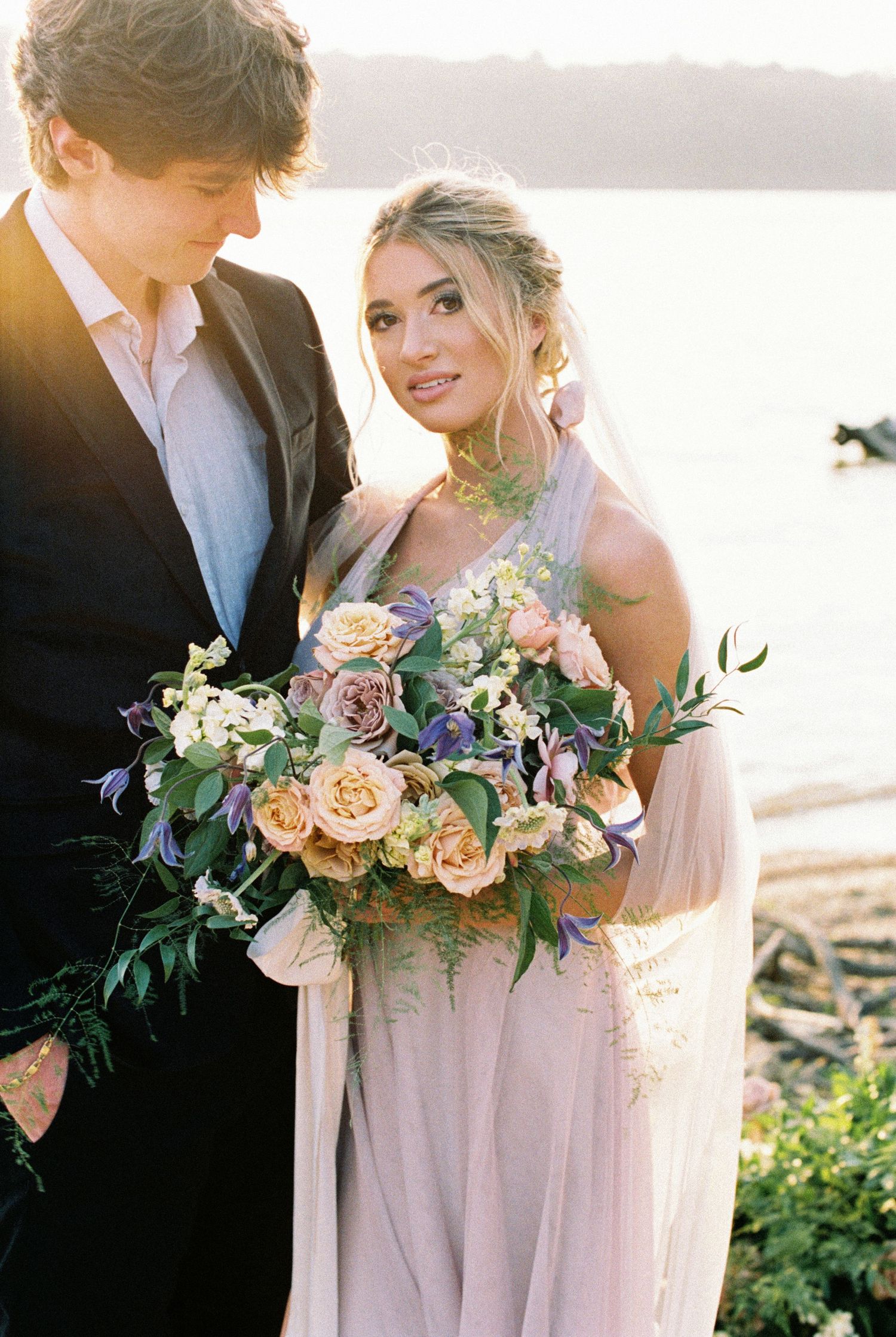Romantic wedding scene with couple holding elegant peach and purple floral bouquet at sunset. Kirstie Whitaker Photography