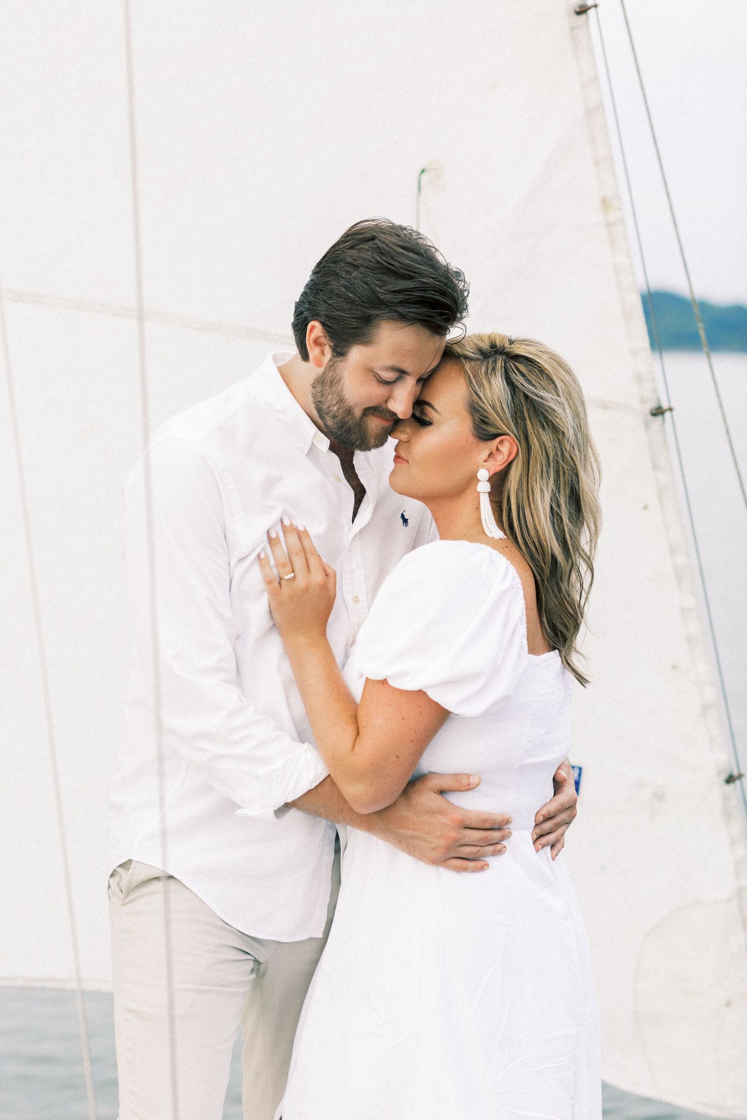 Romantic couple in white outfits sharing an intimate moment against a sailboat backdrop near the ocean.