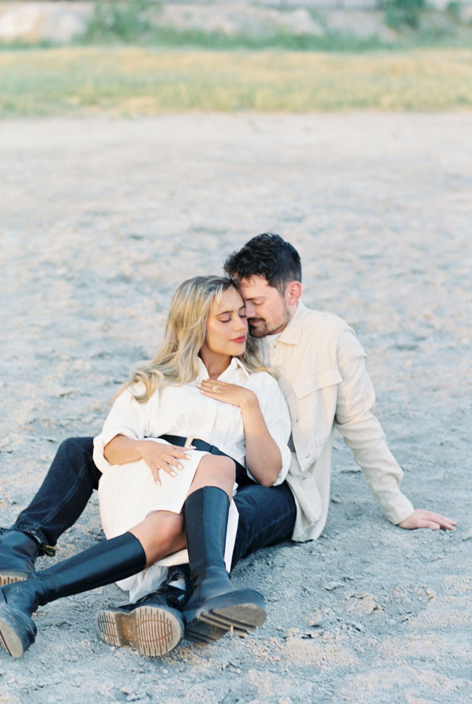 A couple in casual white sweaters and jeans sits intimately together on the sandy beach during a peaceful moment.