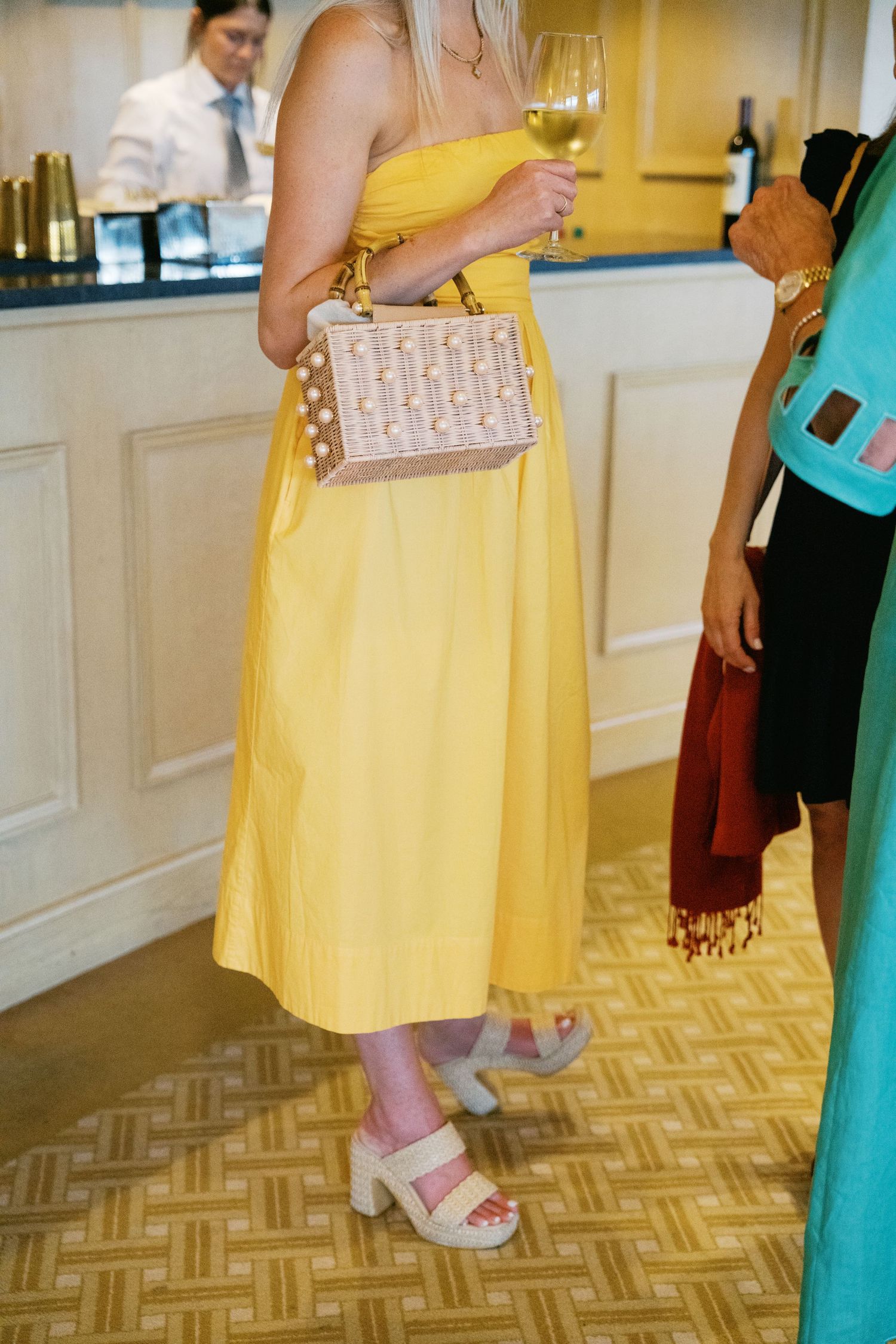 Person in a yellow sundress and pink sandals holds a small clutch purse while standing on a patterned floor.