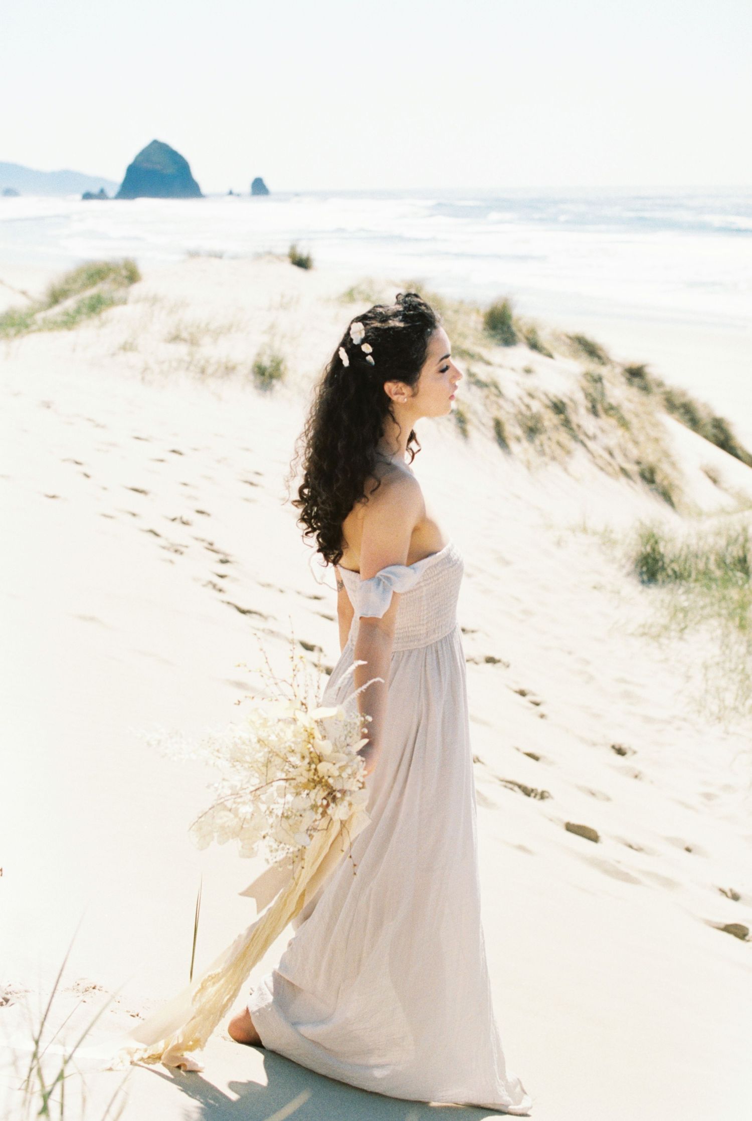 Ethereal beach wedding scene with a flowing off-shoulder dress and dried flowers against Cannon Beach's iconic Haystack Rock.