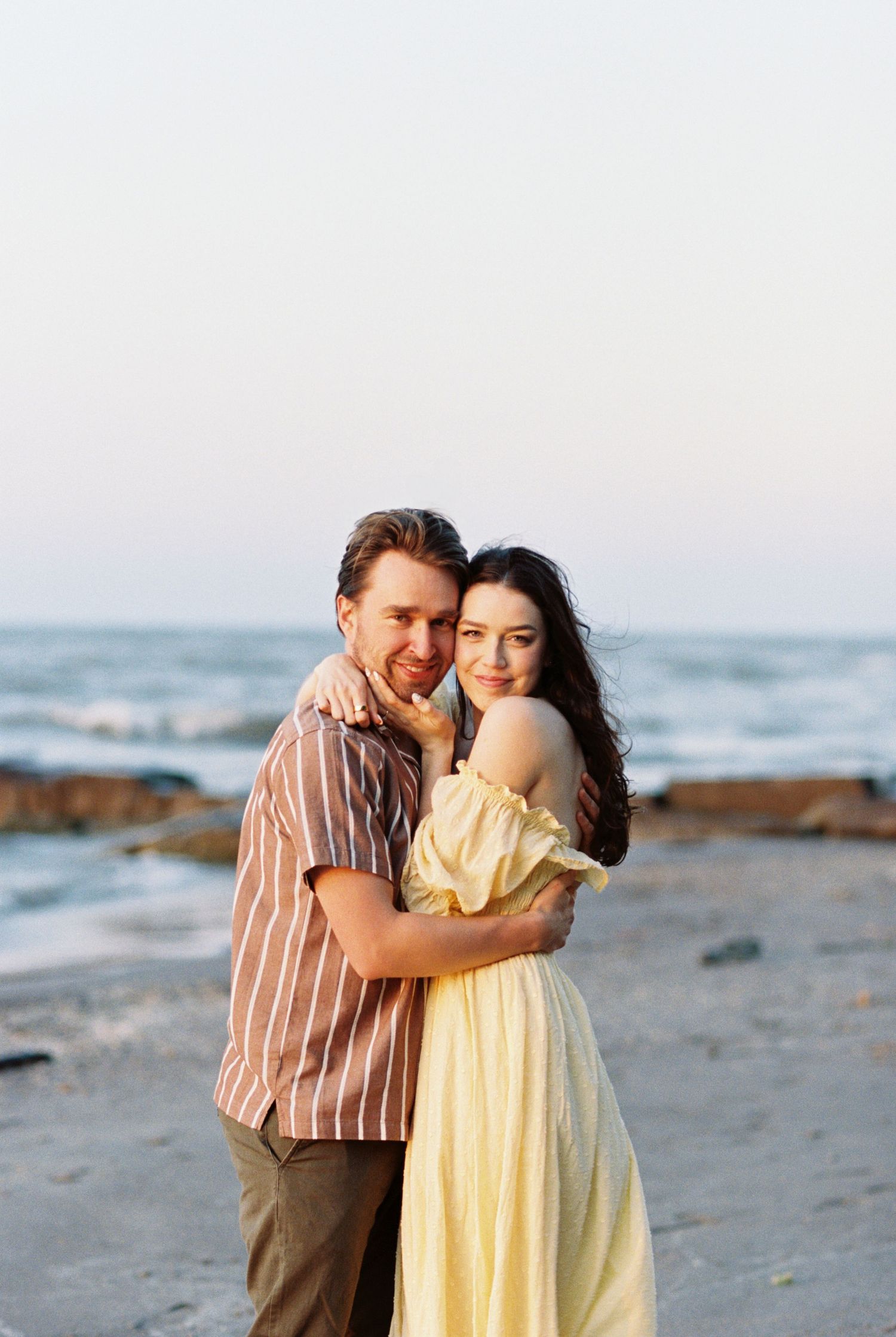 A couple embraces on a rocky beach at sunset with ocean waves in the background.