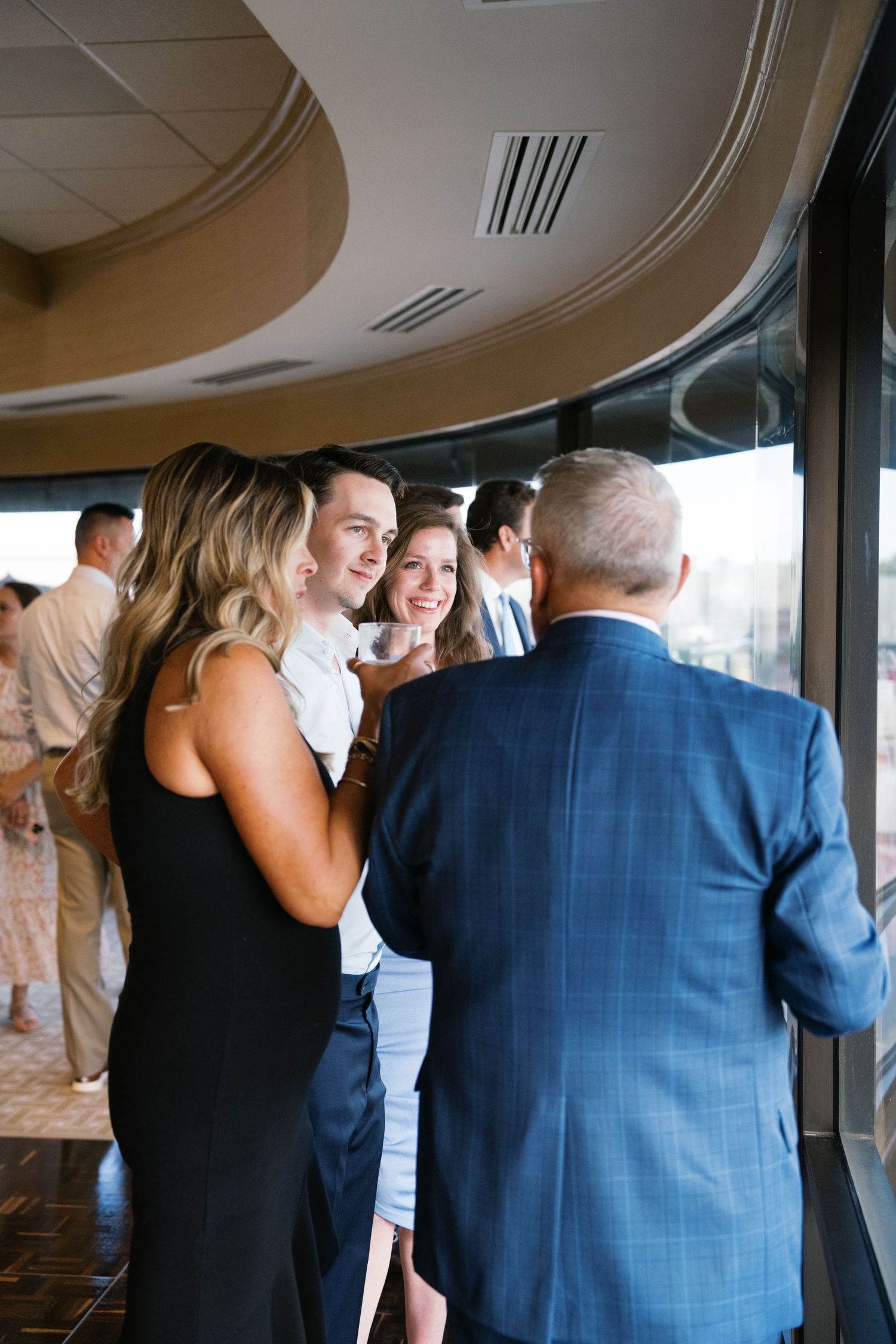 Guests in formal attire gathering by large windows at an event with curved ceiling details.