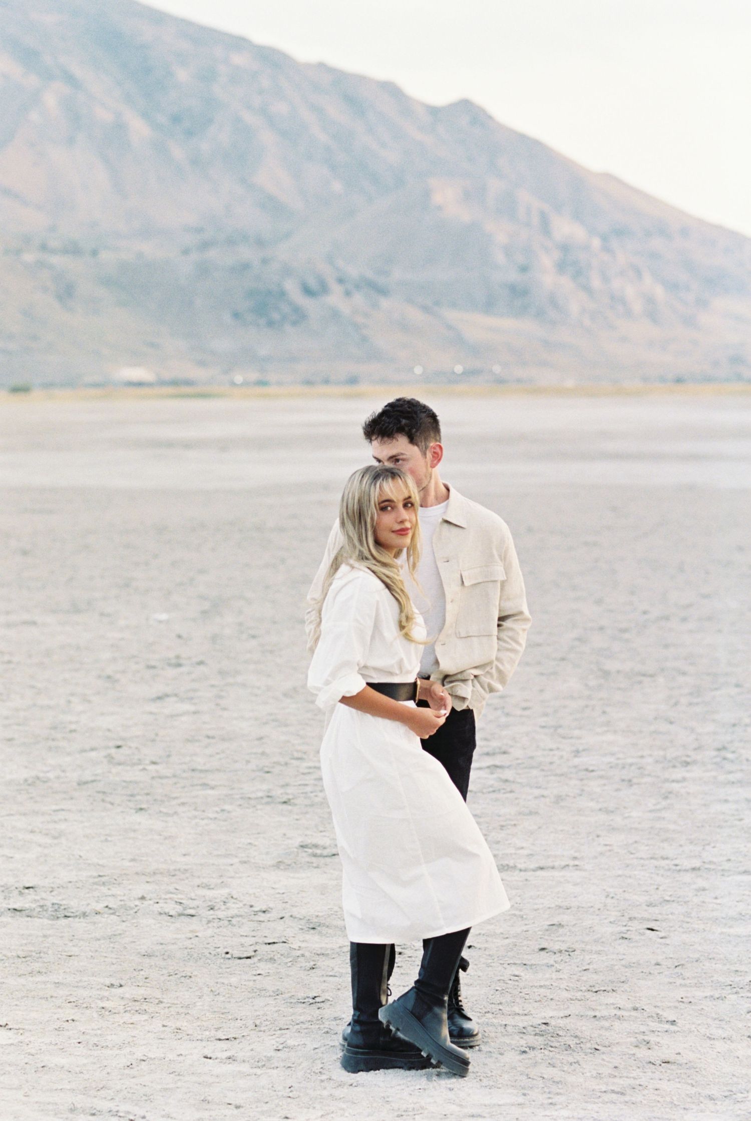 A couple embraces on a desert plain with mountains in the background while wearing white outfits and black boots.