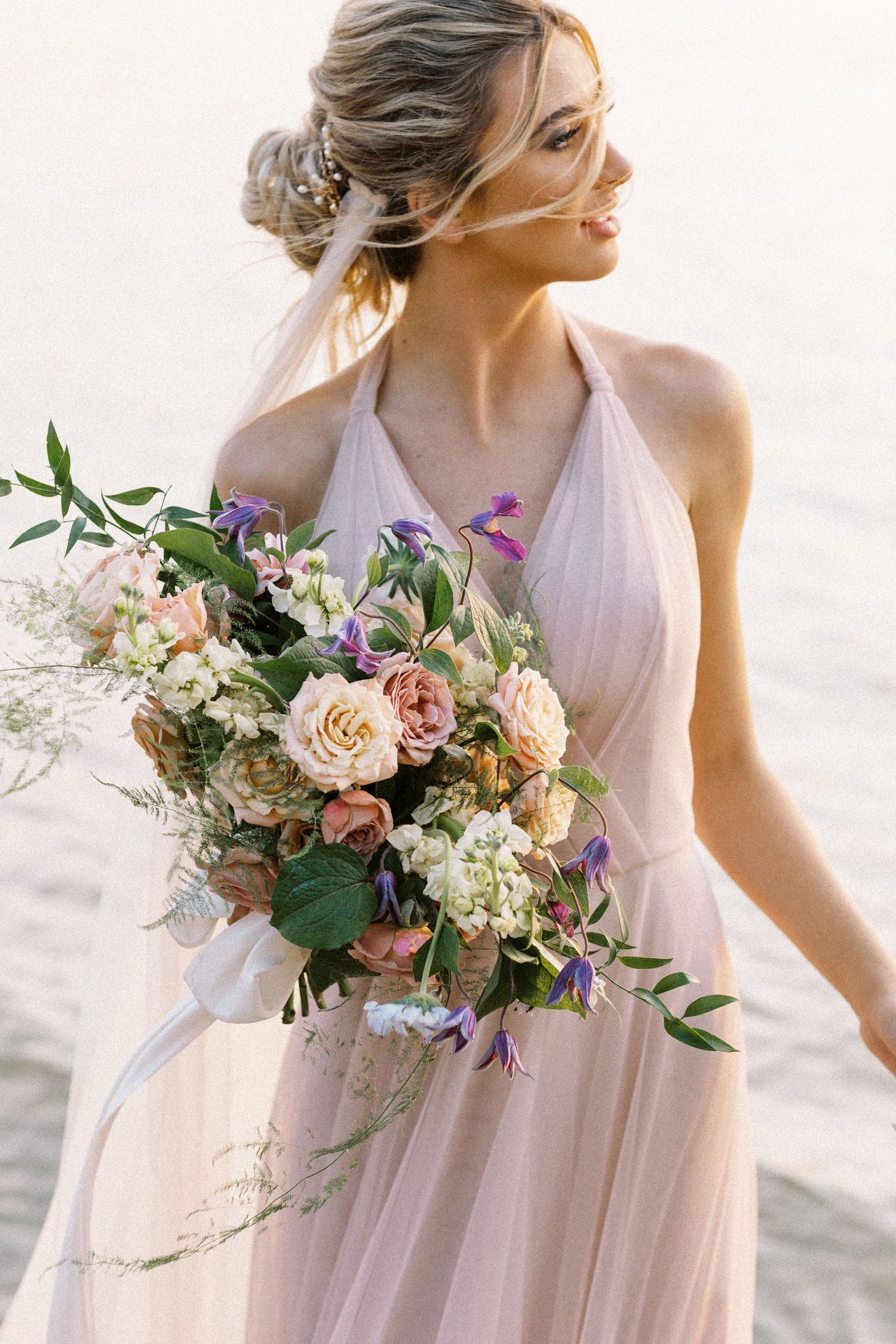 Ethereal bride in flowing blush gown holds romantic bouquet with pink roses and purple wildflowers on beach.