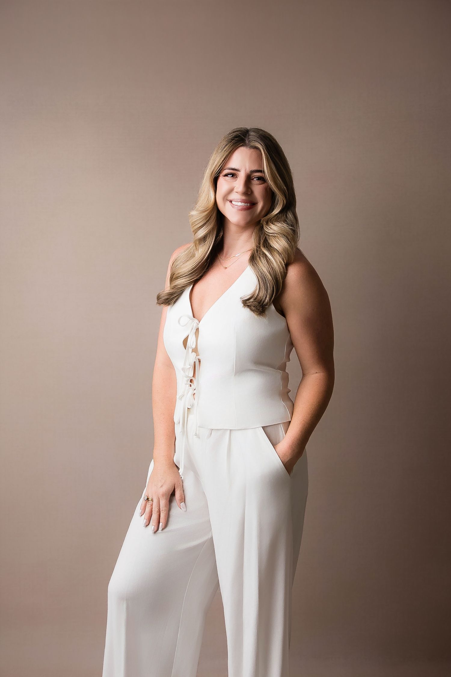 A person wearing an elegant white sleeveless jumpsuit poses against a neutral beige background in a studio setting.