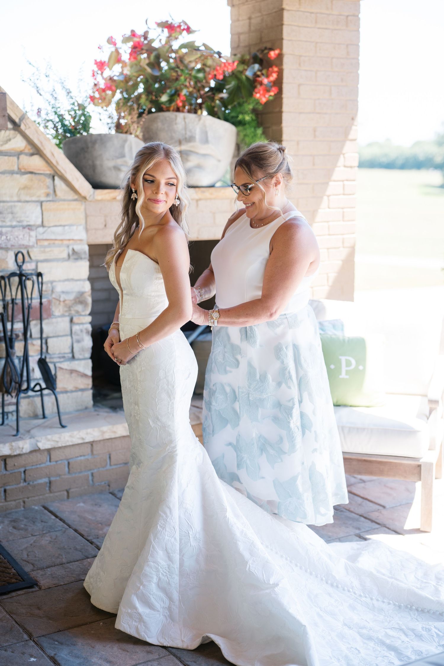 A wedding dress fitting scene shows a helper adjusting the gown's train on an outdoor patio with pink flowers in background.