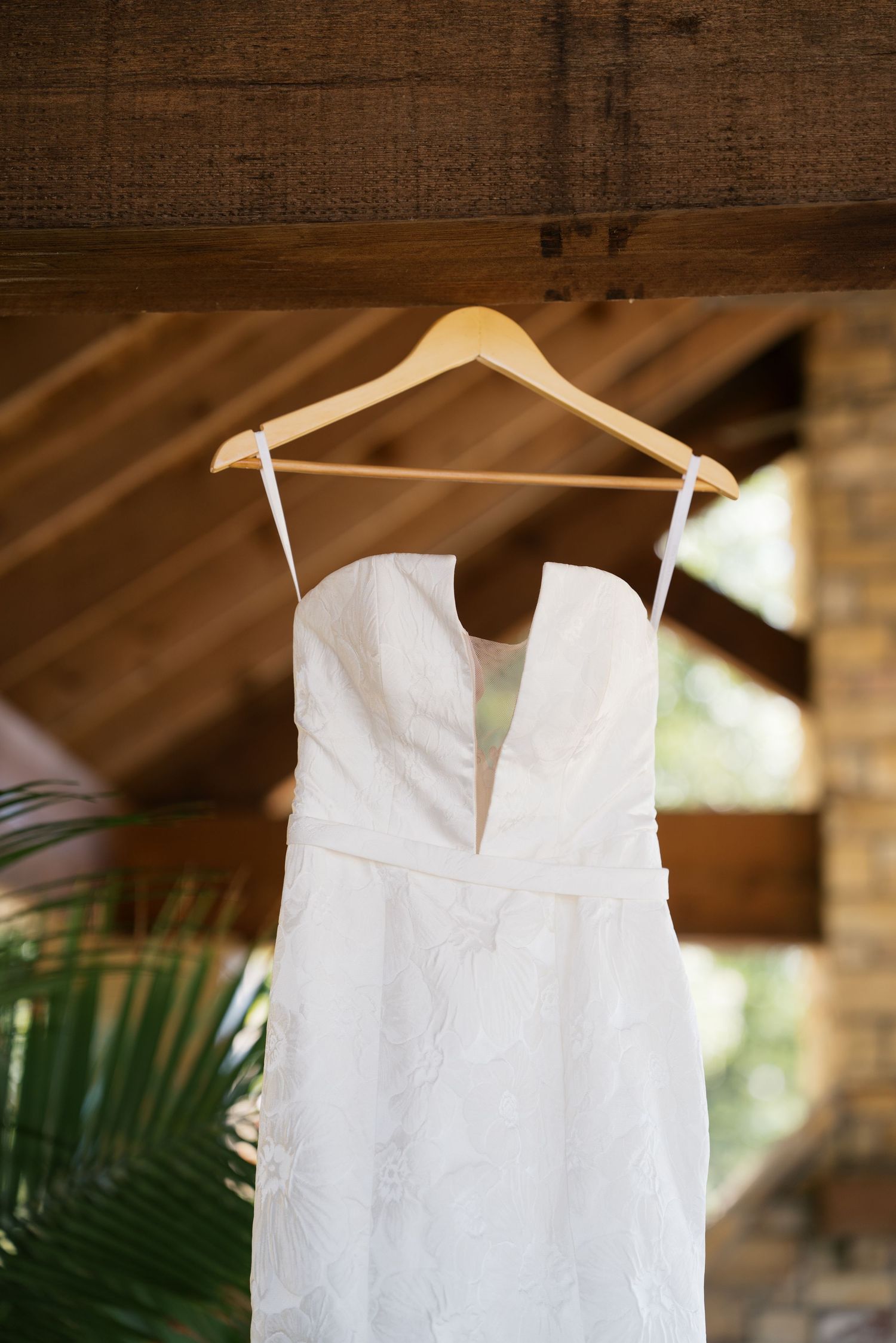 A white wedding dress hangs on a wooden hanger against a rustic backdrop with palm fronds visible in the background.